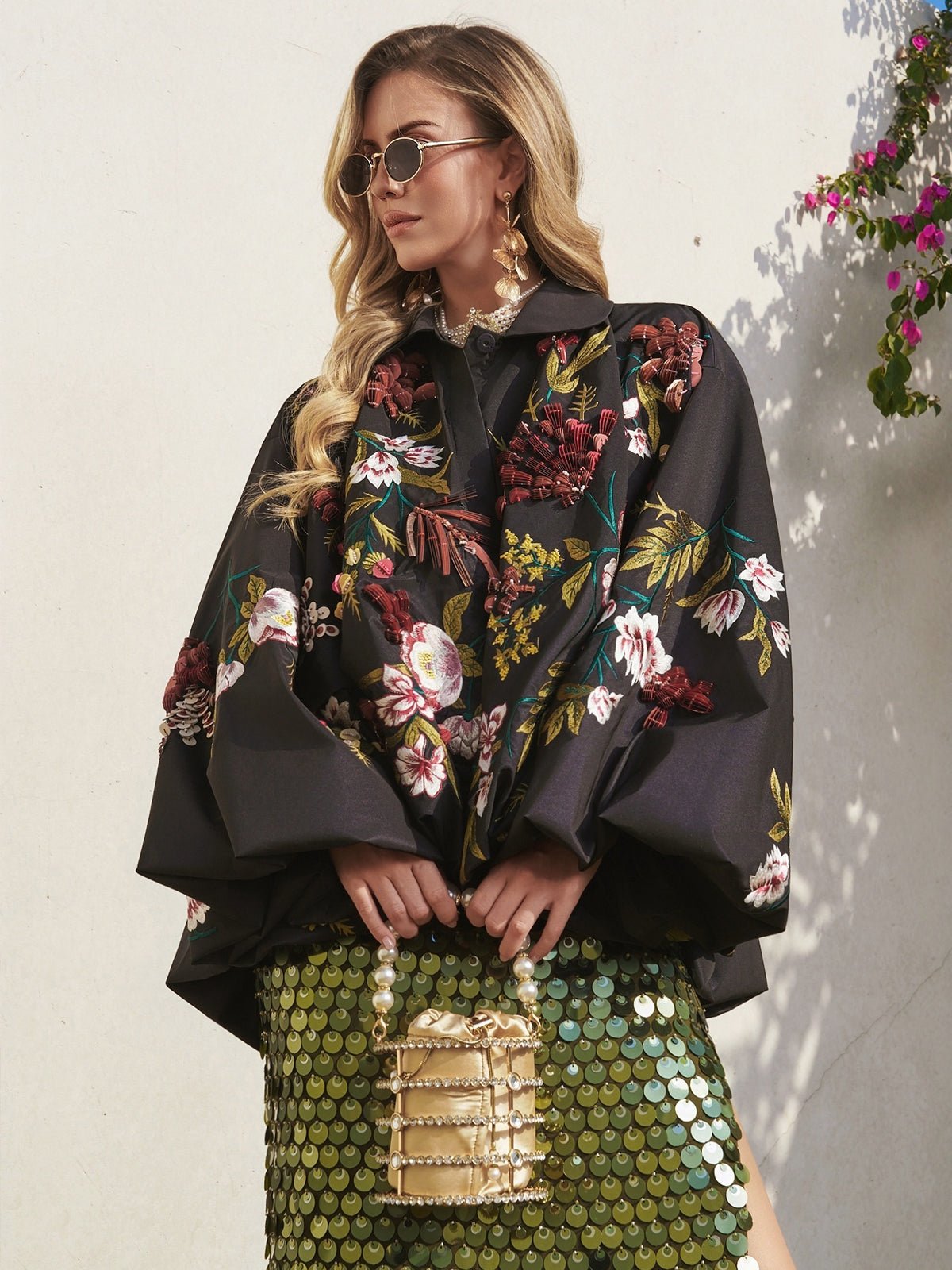 A woman in Social Queen’s Floral Embroidered Floral Puff Sleeve Jacket with a cape silhouette, shiny green skirt, and gold purse stands by a light wall with flowers—an ideal stylish wedding guest look.