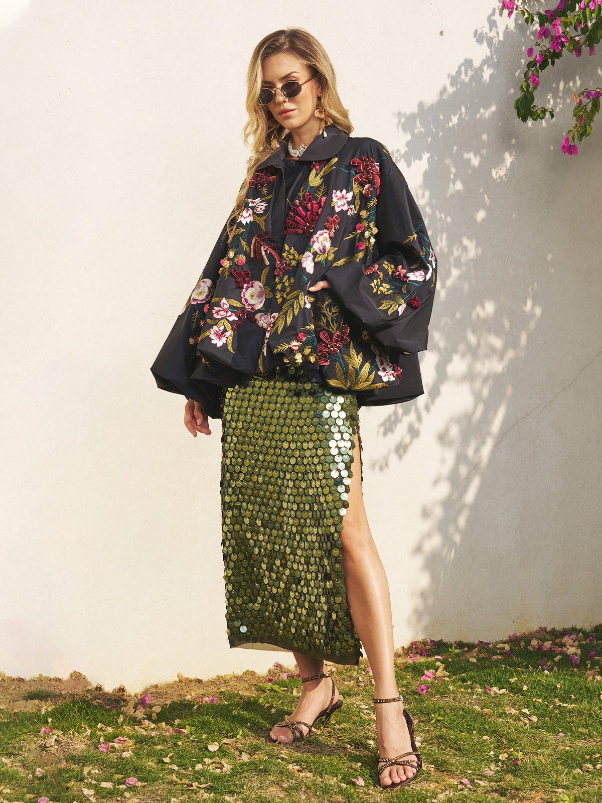 A woman stands outdoors against a white wall, wearing sunglasses, a black floral top, a green sequin skirt with a thigh-high slit, and layered with the Social Queen Floral Embroidered Floral Puff Sleeve Jacket for an elevated wedding guest look.