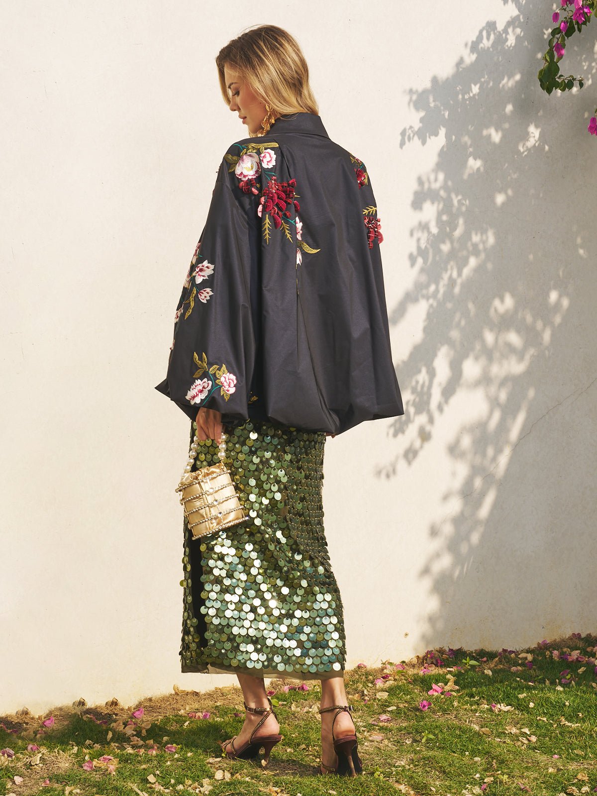 A woman stands on grass wearing the Social Queen Floral Embroidered Floral Puff Sleeve Jacket, a green sequin skirt, heeled sandals, and holding a woven handbag—an elegant wedding guest look. Flowers and shadows line the white wall behind her.