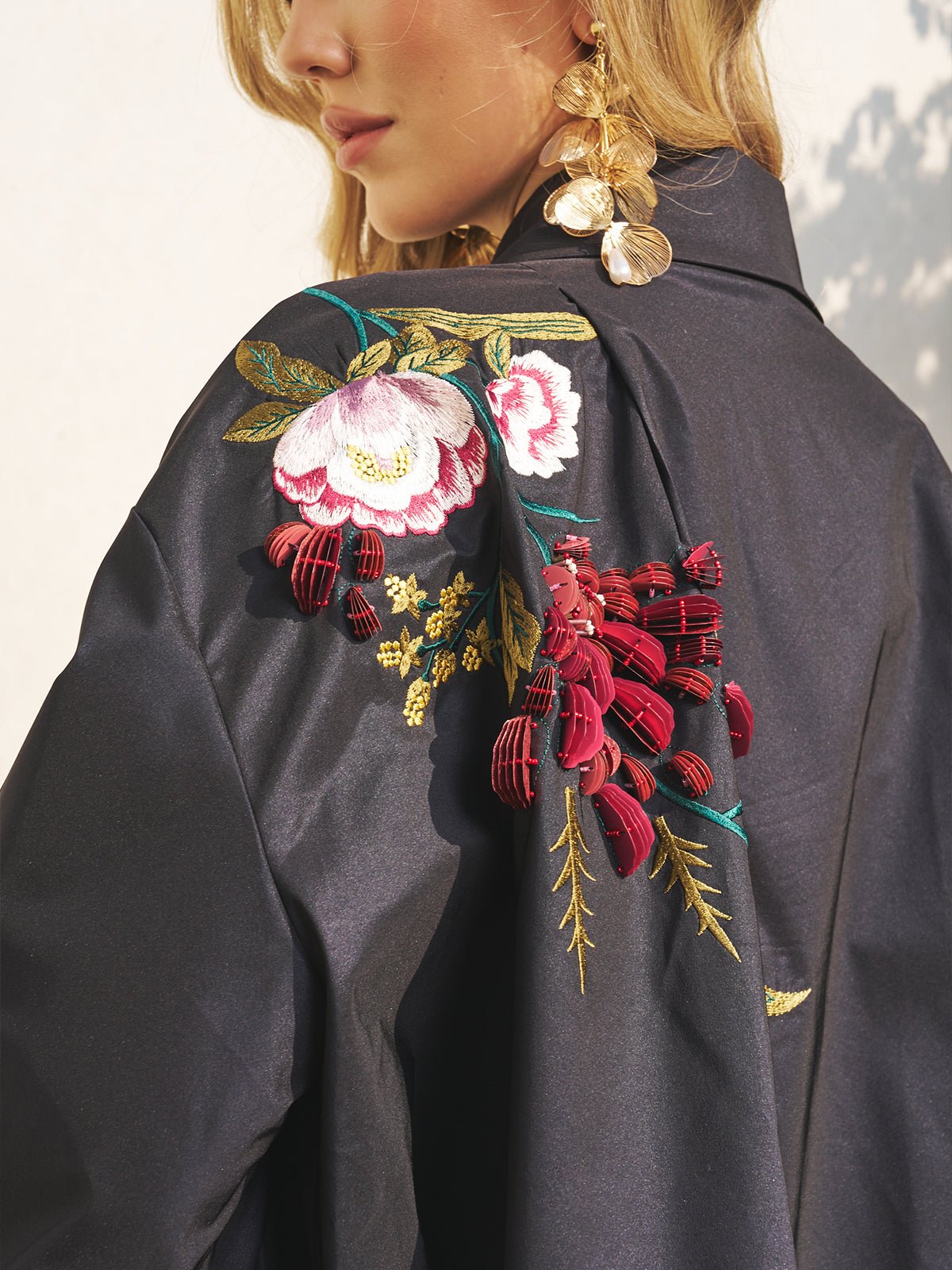 A woman wears the Social Queen Floral Embroidered Puff Sleeve Jacket with bold floral designs and red accents, gold leaf earrings, and loose blonde hair—an ideal, stylish look for a wedding guest.