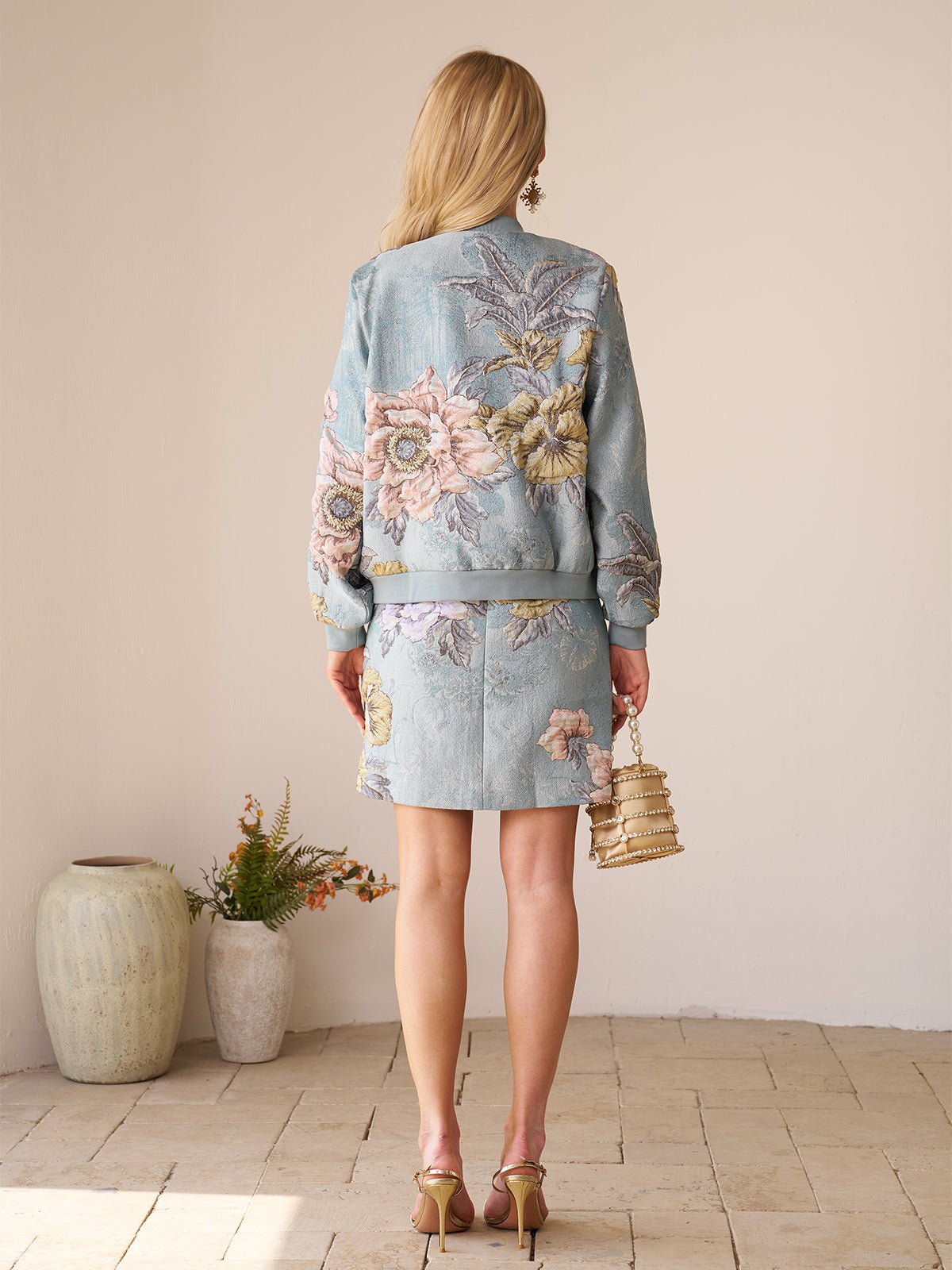 A woman with long blonde hair stands indoors on a tiled floor near potted plants, wearing the Social Queen Floral Embroidered Jacquard Jacket and holding a small handbag.