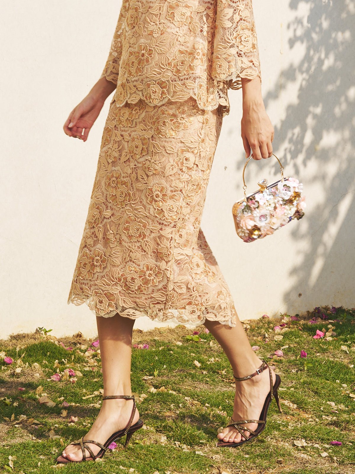 A person walks outdoors on grass with flower petals, wearing Social Queen’s Floral Embroidered Lace Midi Skirt, a matching top, strappy sandals, and carrying a small floral handbag.