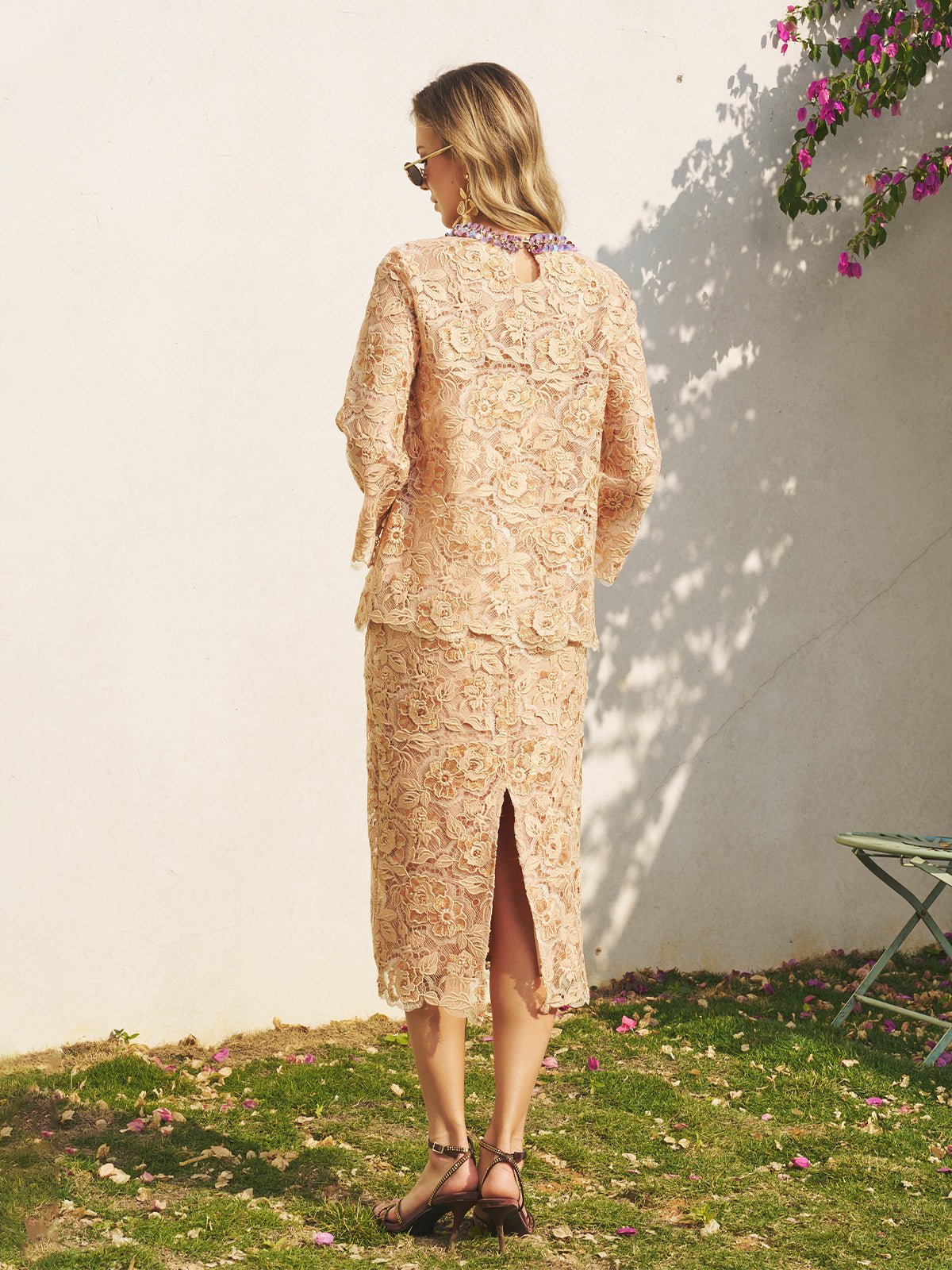 A woman, facing away outdoors on grass by a white wall and flowers, wears the Social Queen Floral Embroidered Lace Top with Beaded Details.