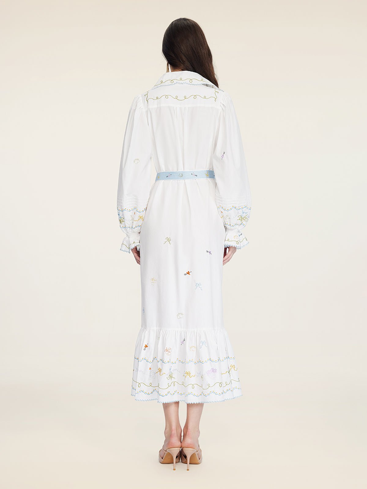 A woman faces away, wearing the Social Queen Floral Embroidered Lantern Sleeve Dress, featuring hand-embroidered floral motifs, a light blue belt, and beige heeled sandals.
