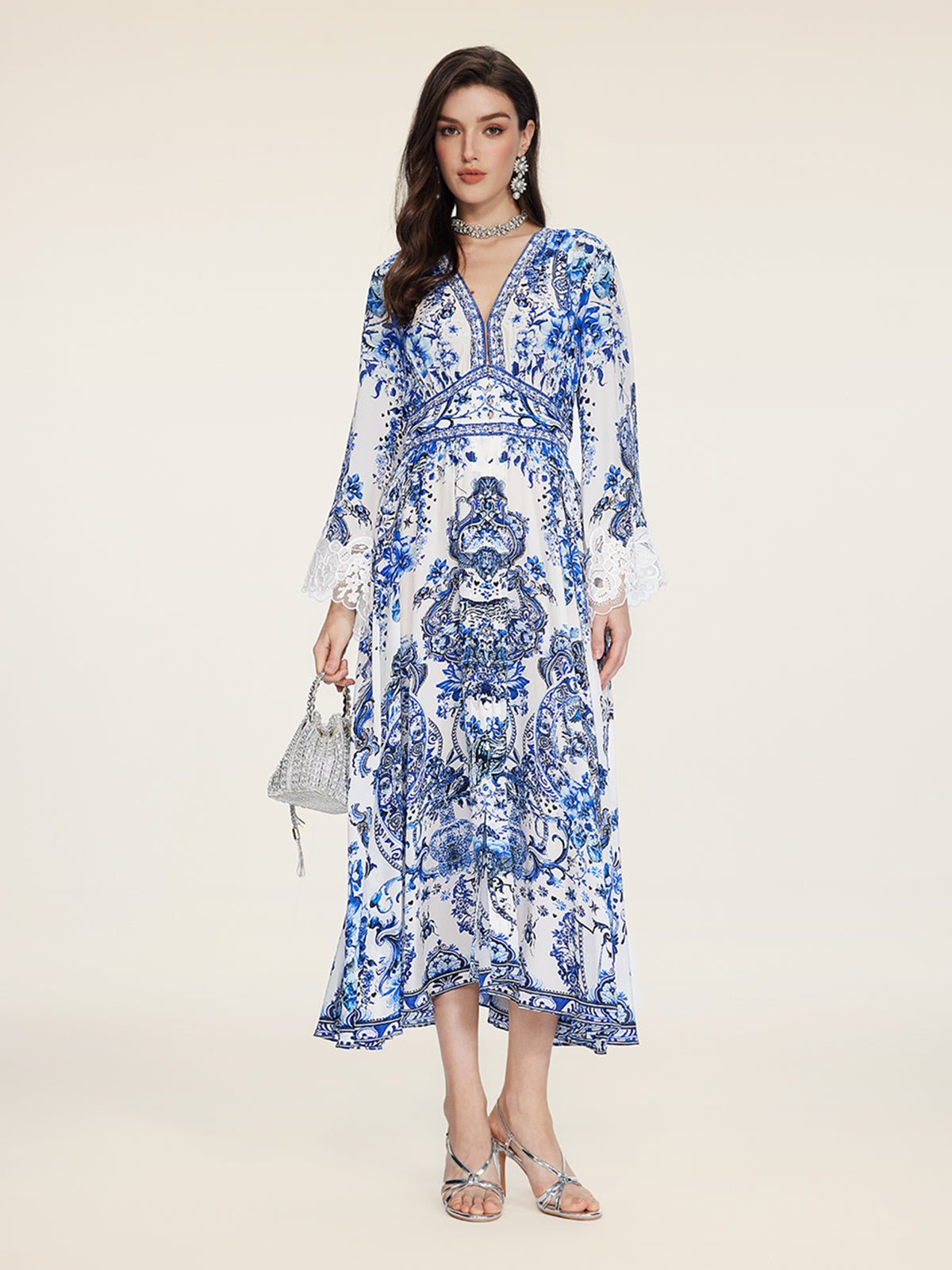 A woman stands against a plain background in the Social Queen Floral Embroidered Lantern Sleeve Dress, featuring white and blue floral patterns, a deep V-neck, and long sleeves. She holds a small silver handbag and wears silver sandals with jewelry.