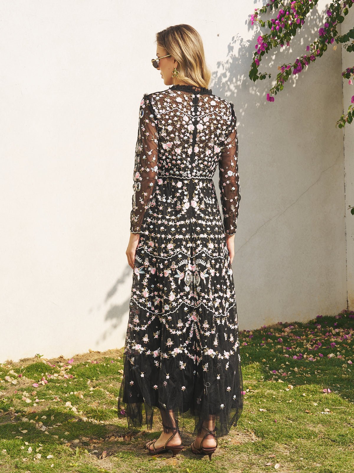 A woman stands on grass facing a white wall, wearing the Social Queen Floral Embroidered Mesh Dress with sheer panels and sandals—a chic choice for a wedding guest.