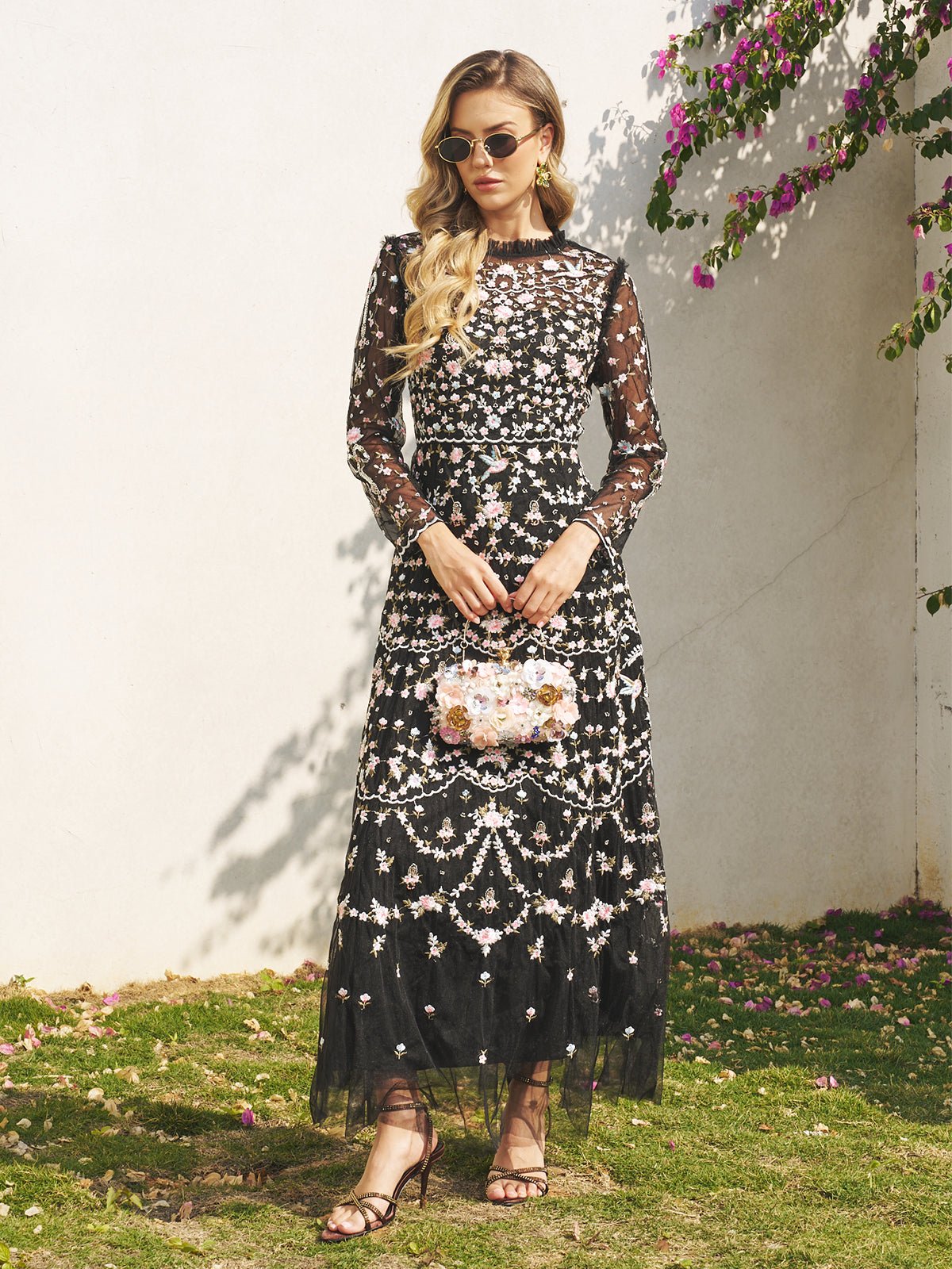 A woman wearing sunglasses showcases the Social Queen Floral Embroidered Mesh Dress—long-sleeved with floral details—as she stands on grass before a white wall, holding a matching floral handbag. Perfect as a wedding guest dress.