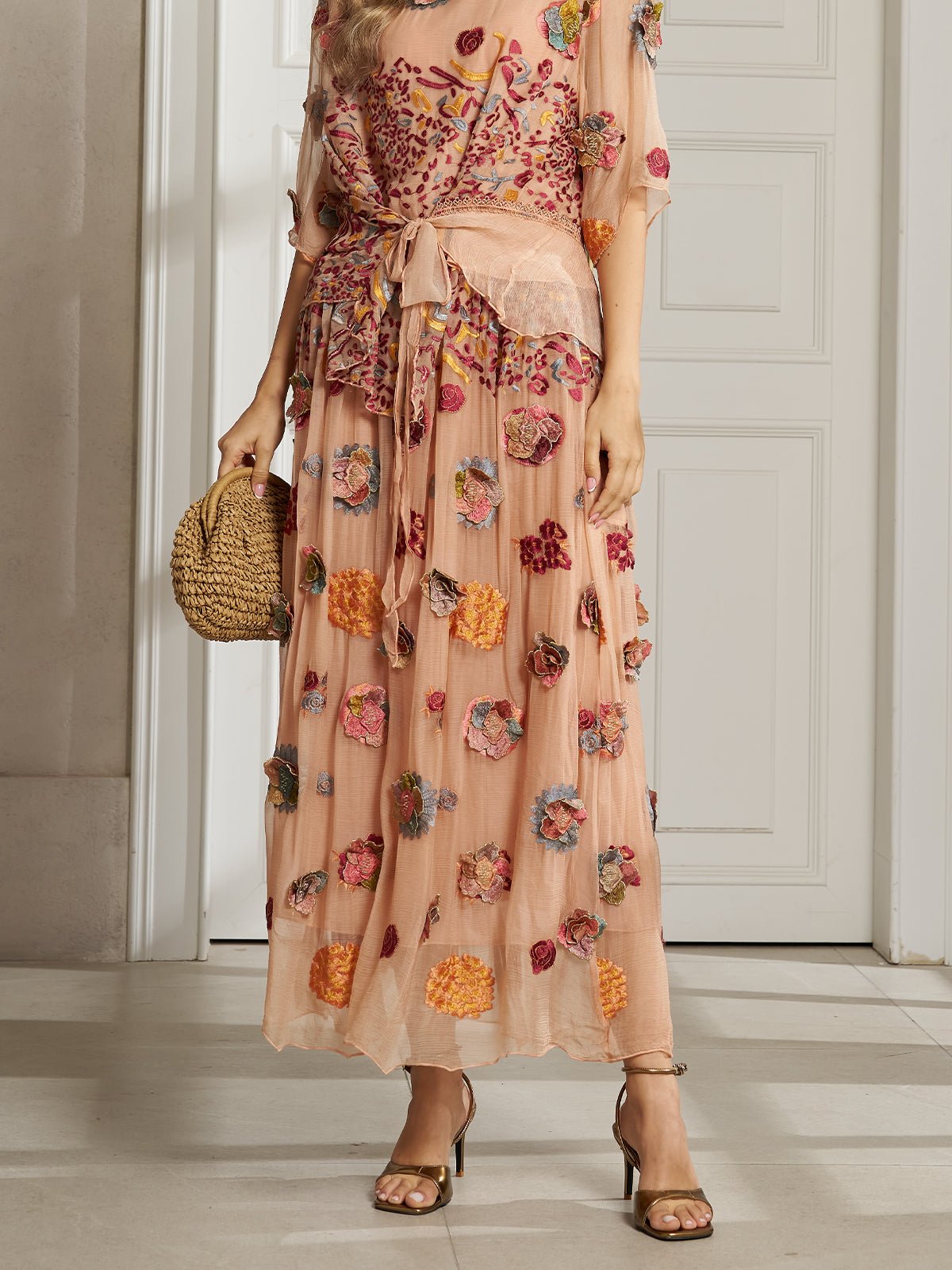 A woman stands before a white paneled door in a sheer peach dress with a waist tie, carrying a woven bag. She completes her look with the Social Queen Floral Embroidered Silk A-Line Skirt for subtle elegance.