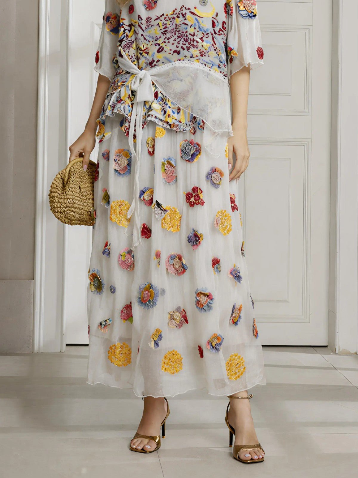 Indoors, a person wears Social Queen's Floral Embroidered Silk A-Line Skirt with a matching tied top, beige heels, and holds a woven bag.
