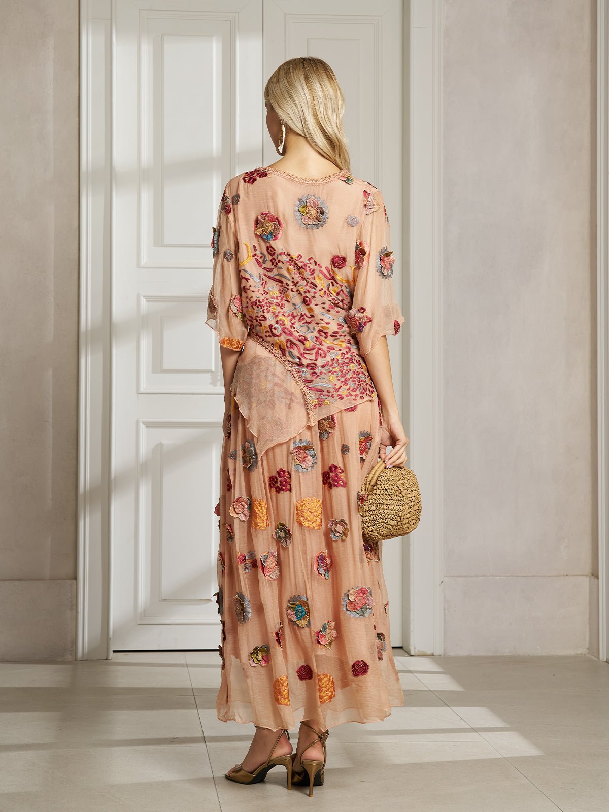 A woman stands indoors by a white door, wearing the Social Queen Floral Embroidered Silk A-Line Skirt with heels and holding a small woven handbag.