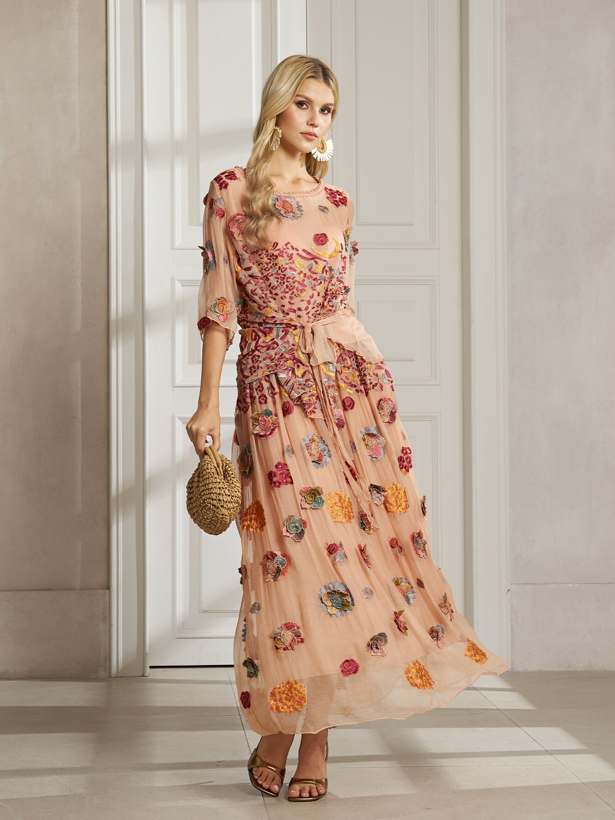 A woman stands indoors wearing the Social Queen Floral Embroidered Silk A-Line Skirt, paired with a sheer peach dress, woven handbag, and sandals.