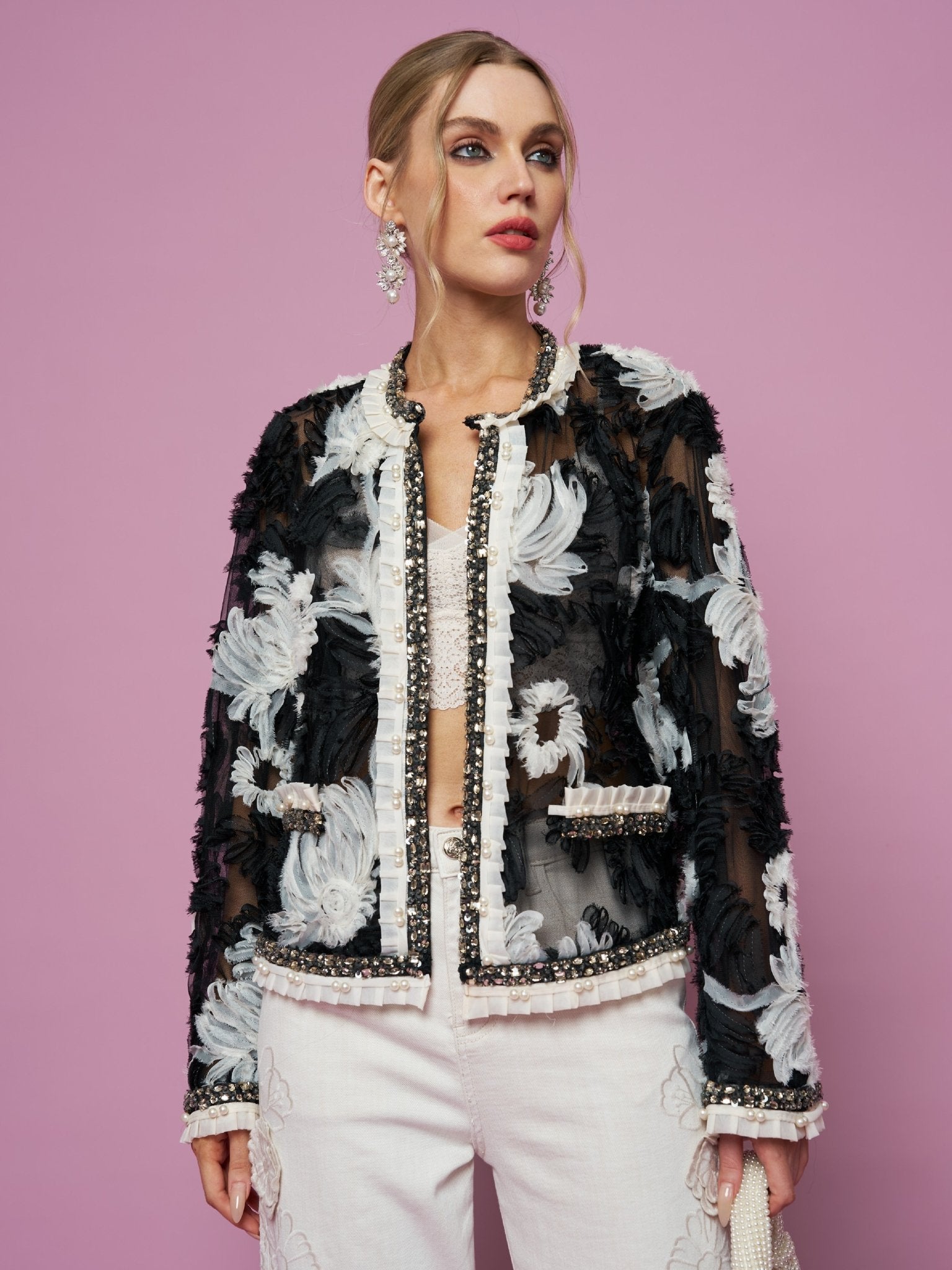 A woman poses against a pink backdrop wearing the Social Queen Floral Embroidered Tulle Beaded Jacket, paired with white pants and statement earrings, holding a bag in her left hand.