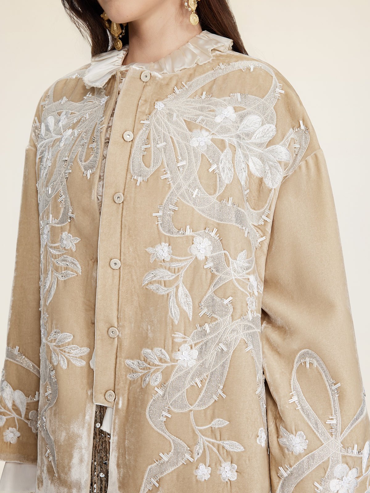 A person wears Social Queen’s Floral Embroidered Velvet Jacket in beige with hand-embroidered floral motifs and lace details; gold earrings and a white collar are also visible.