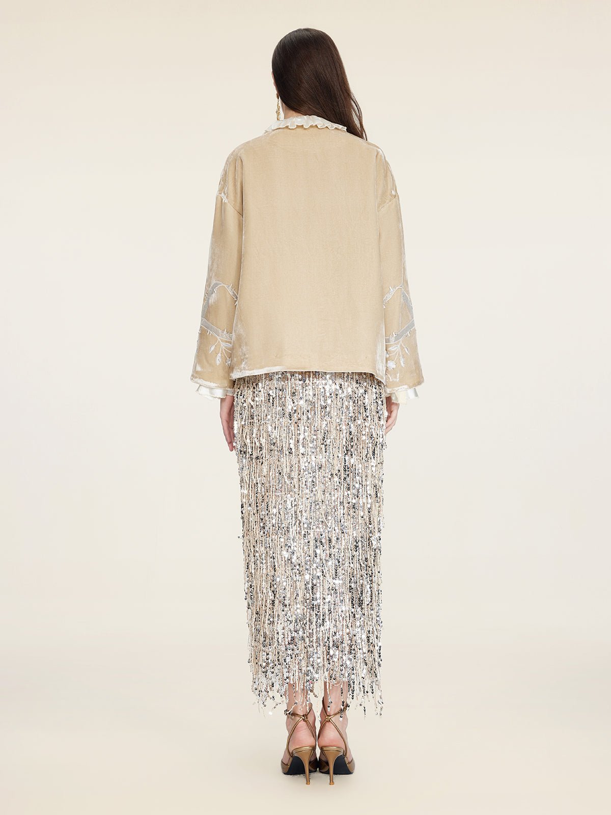 A woman faces away wearing Social Queen's Floral Embroidered Velvet Jacket and a long silver fringe skirt with black strappy heels, set against a plain light background.
