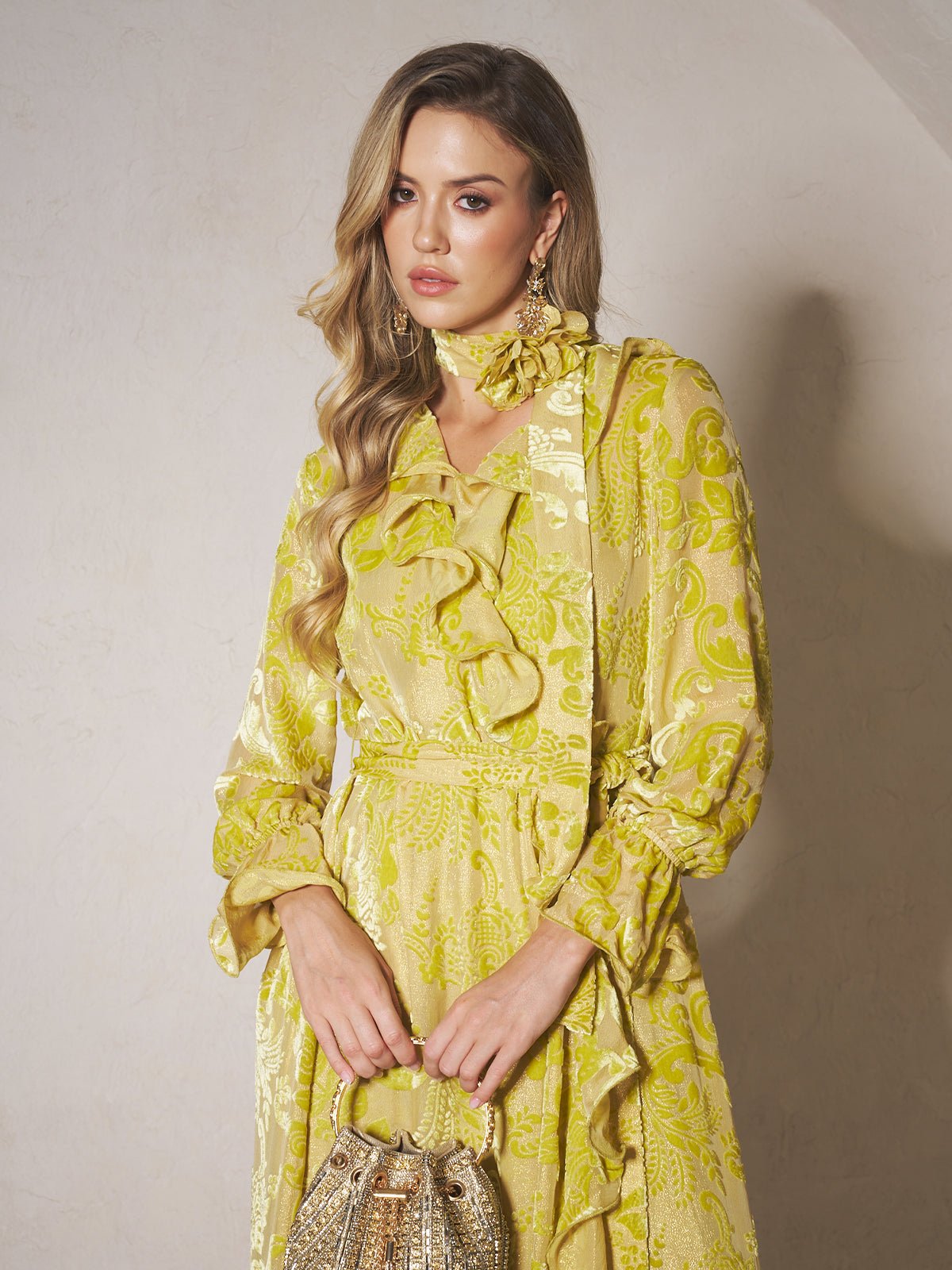 A woman in the Social Queen Floral Jacquard Velvet Ruffles Dress, featuring long sleeves and yellow floral patterns, holds a gold handbag while standing against a light-colored wall.