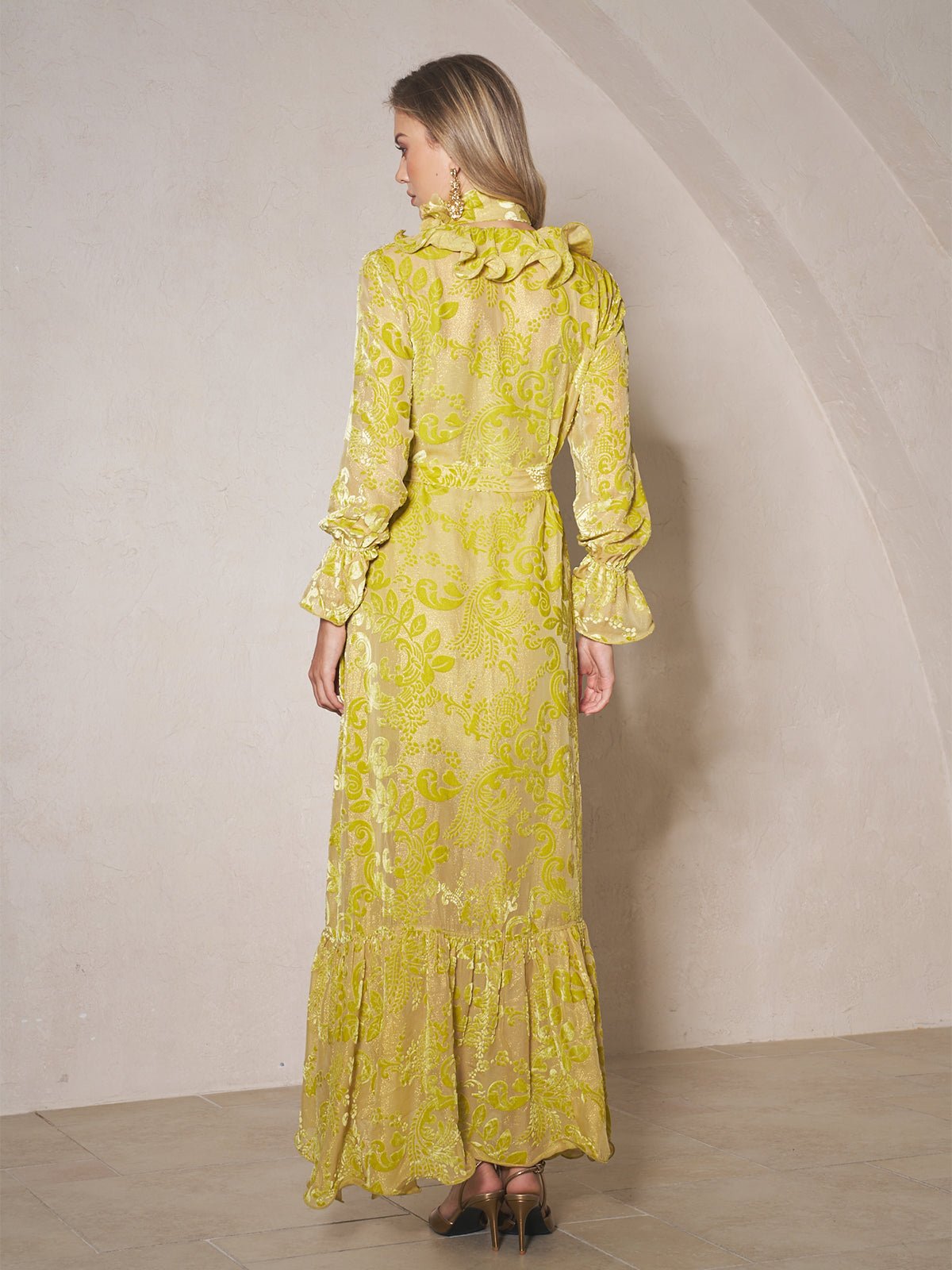 A woman stands indoors with her back to the camera, wearing the Social Queen Floral Jacquard Velvet Ruffles Dress—a long wedding guest dress with ruffled details, long sleeves, and a wrap silhouette—paired with high-heeled shoes.