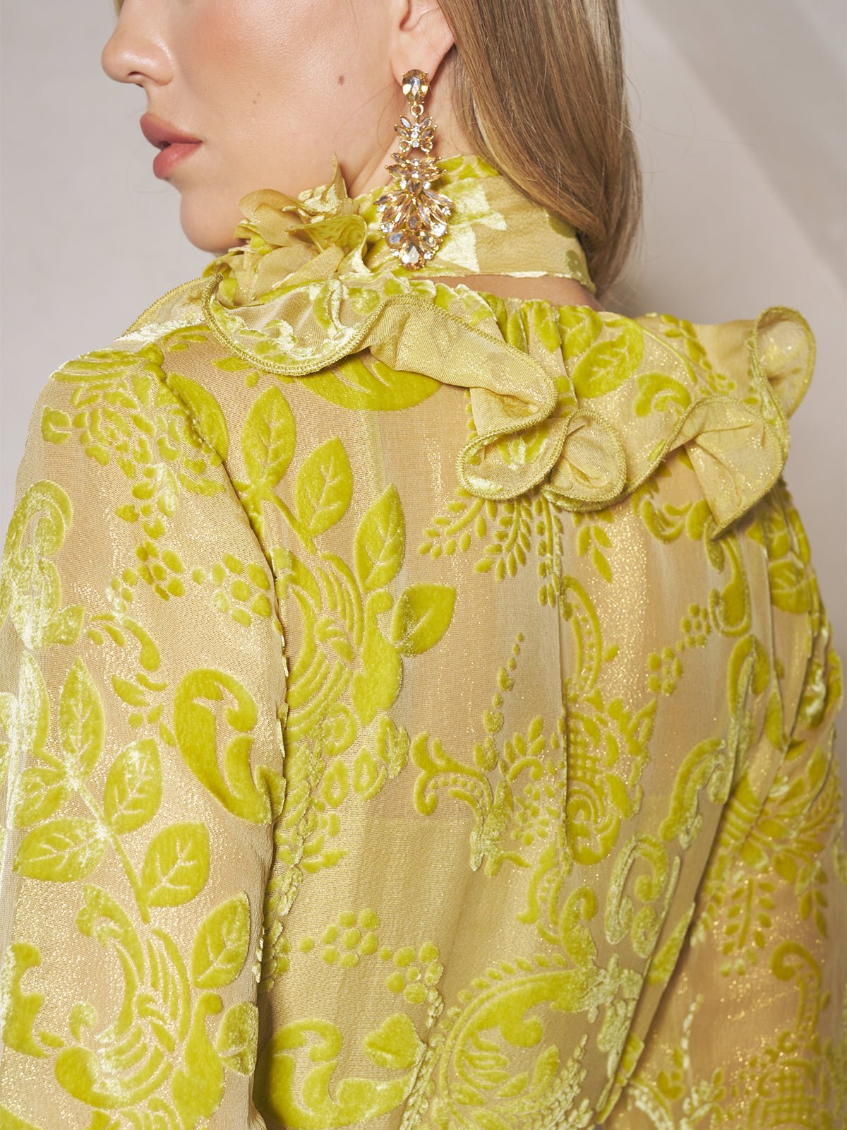 A person, shown from the back and side, wears Social Queen's Floral Jacquard Velvet Ruffles Dress, featuring a sheer yellow-green floral pattern with ruffles and ornate gold earrings.