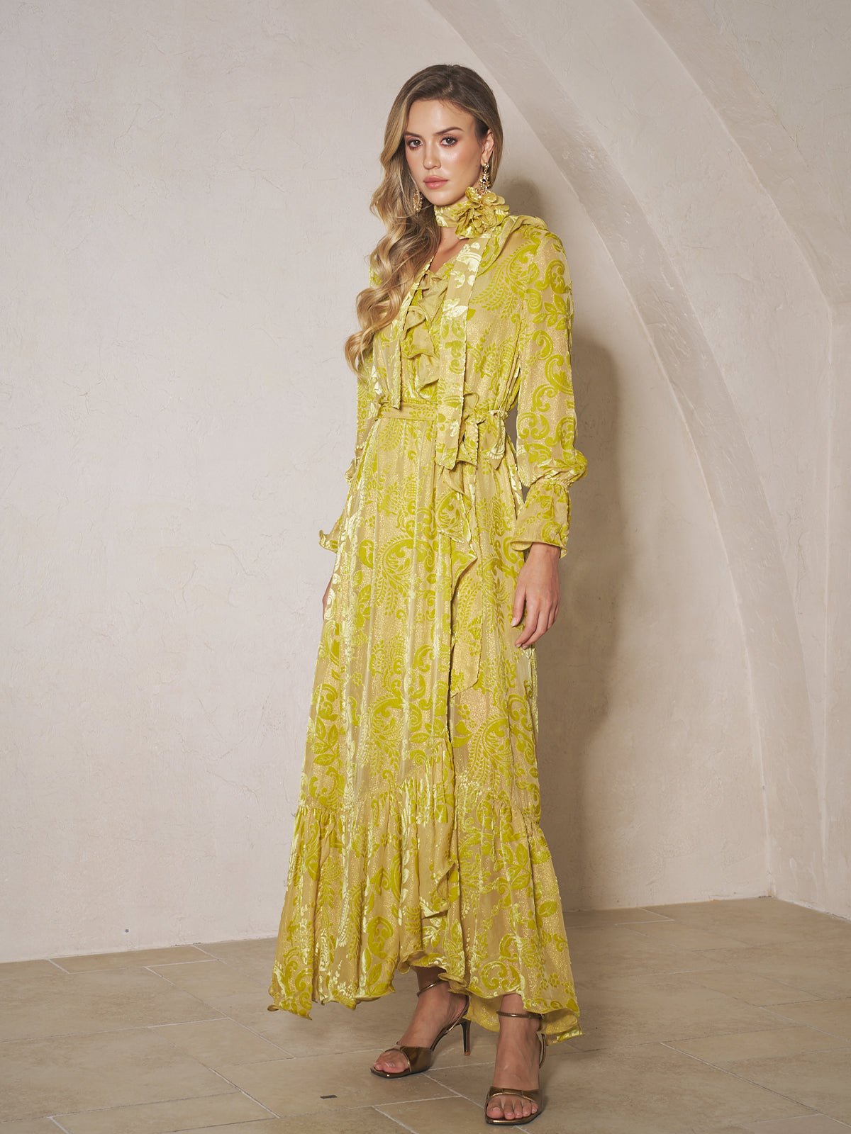 A woman stands indoors wearing the Social Queen Floral Jacquard Velvet Ruffles Dress with long sleeves, ruffles, and a tie at the neck—an elegant choice for wedding guests. She has long wavy hair and heeled sandals.