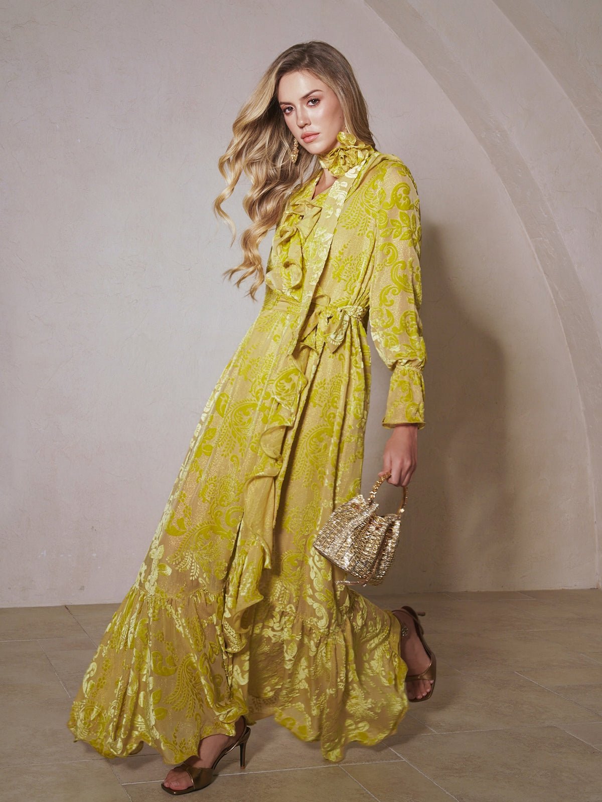 A woman stands indoors wearing the Social Queen Floral Jacquard Velvet Ruffles Dress in yellow, paired with heeled sandals and a small woven handbag.