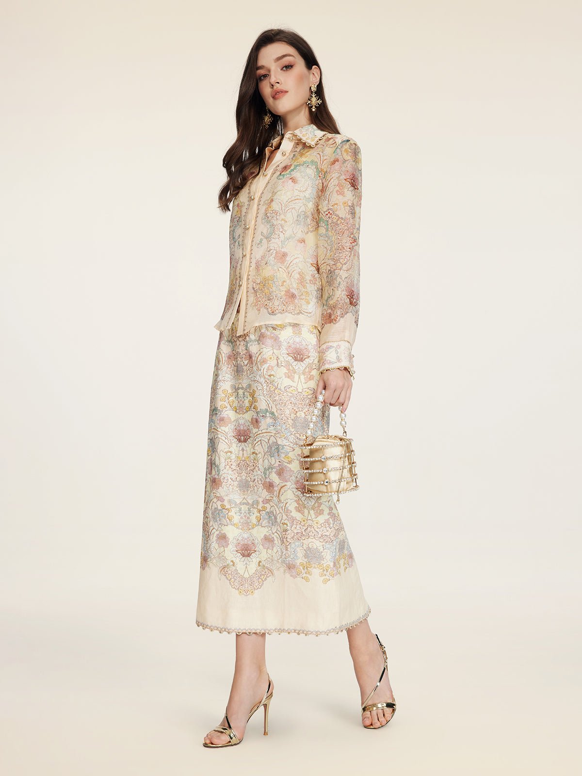 A woman models the Social Queen Floral Print Midi Skirt with a matching blouse, accessorized with a gold handbag and gold high-heeled sandals, set against a plain background.