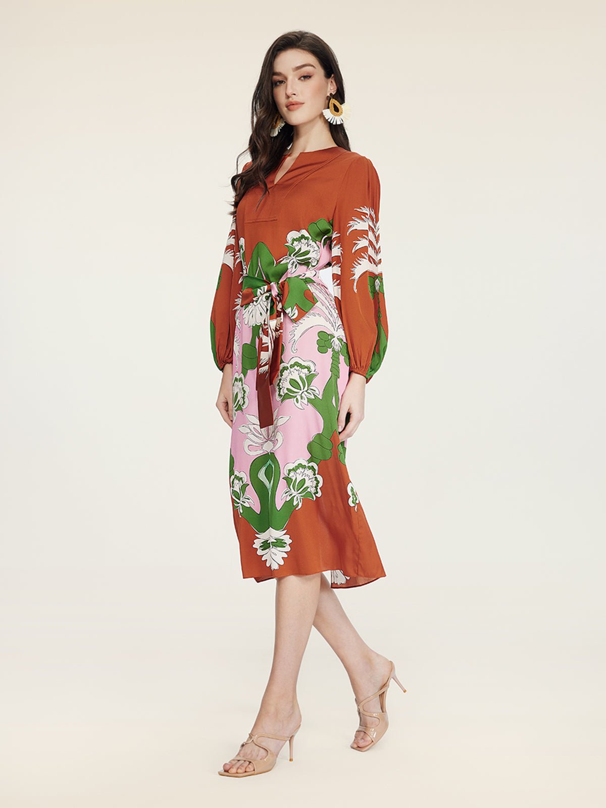 A woman models the Social Queen Floral Printed Silk Dress—rust-colored with long sleeves and green and white florals—paired with nude heeled sandals, exuding vacation elegance against a plain background.