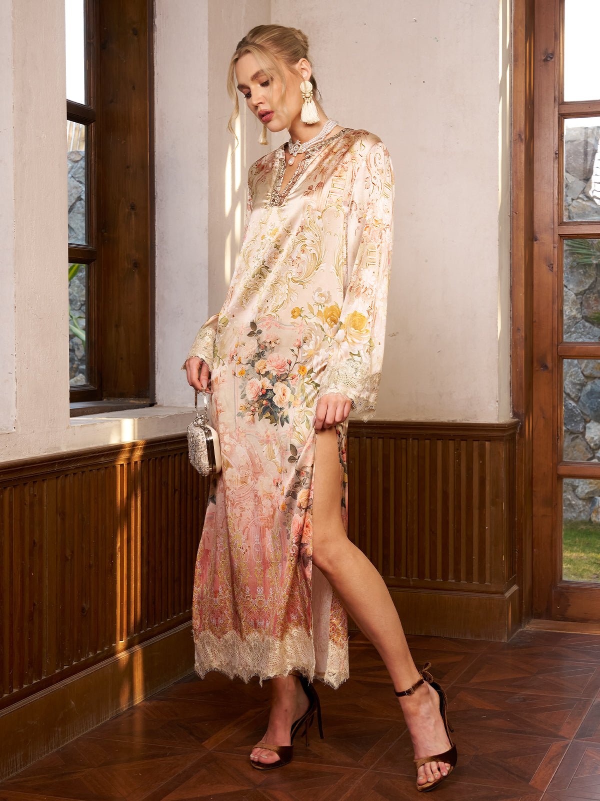 A woman in the Social Queen Garden Print Silk Dress with Lace Trim stands indoors by a window, holding a small purse and wearing black heels and statement earrings—perfect for a wedding guest.