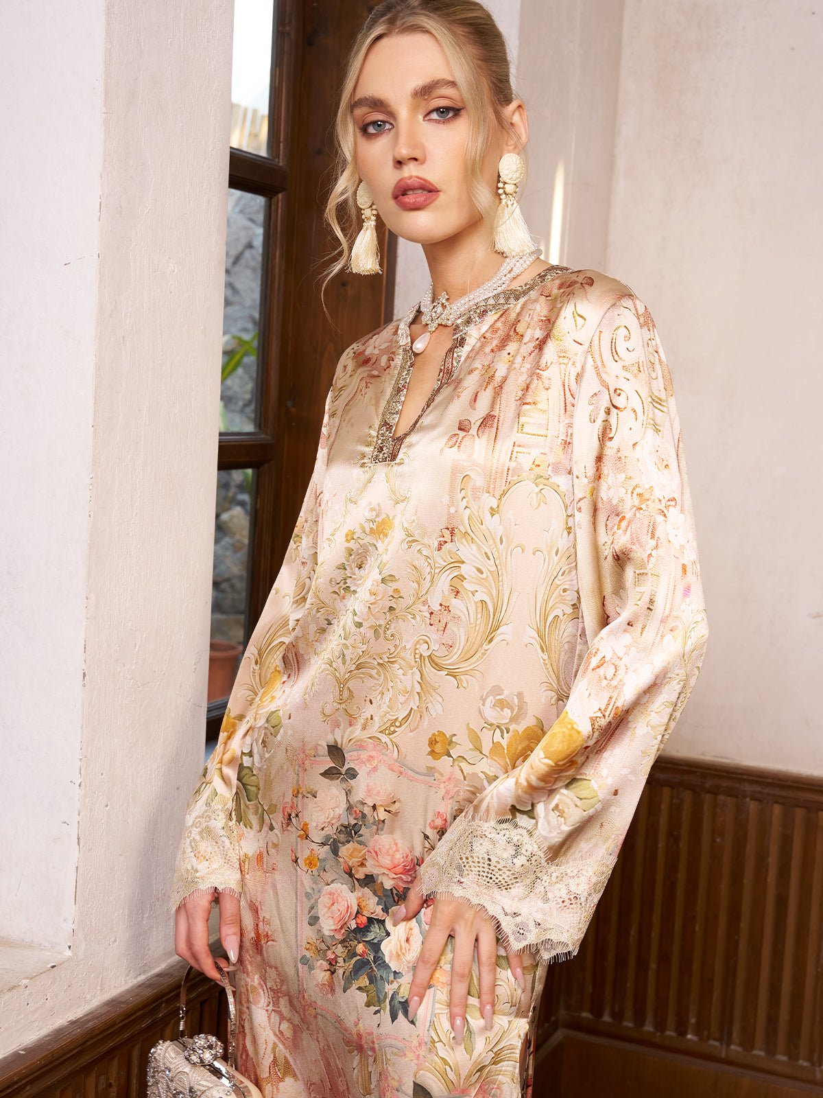 A woman stands by a window in the Social Queen Garden Print Silk Dress with Lace Trim, accessorized with statement earrings, a necklace, and a small clutch—an elegant pick for any wedding guest.