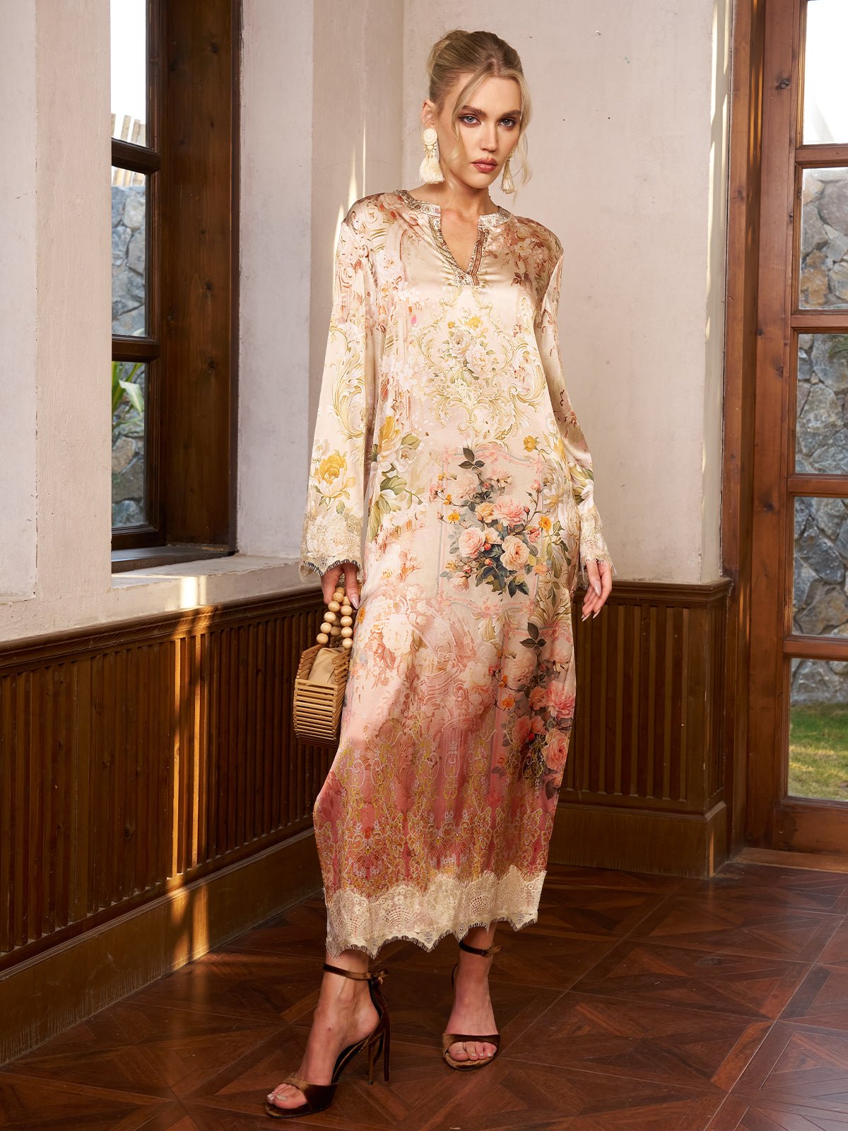 A woman indoors wears the Social Queen Garden Print Silk Dress with Lace Trim, paired with a wicker handbag and strappy heels—an elegant choice for a wedding guest ensemble.