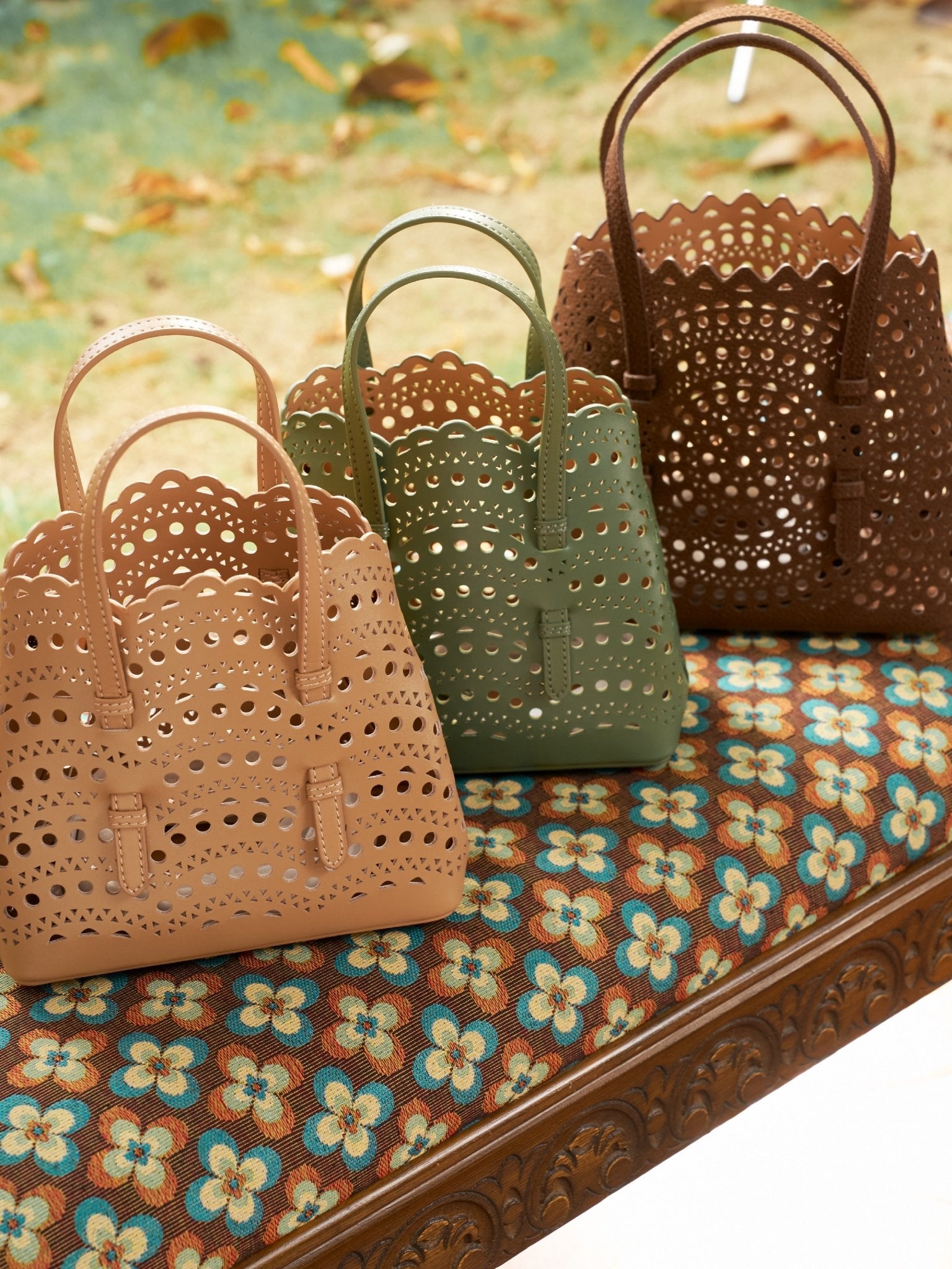 Three Social Queen Geometric Cut-Out Leather Bags in beige, green, and brown are displayed on a floral-patterned bench against a blurred outdoor background.