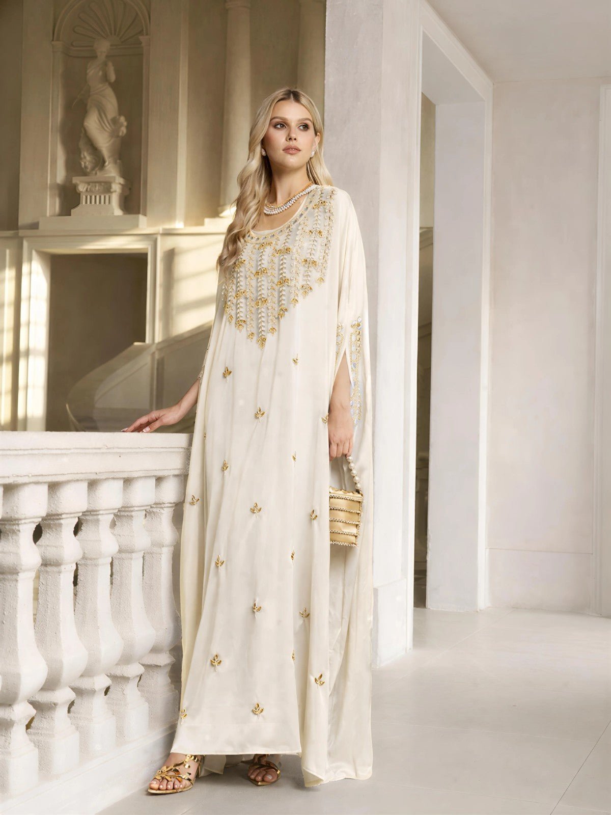 A woman stands indoors by a white railing, holding a small woven handbag and wearing the Gilded Leaf Embellished Cape Dress by Social Queen, surrounded by classical decor and a statue in the background.