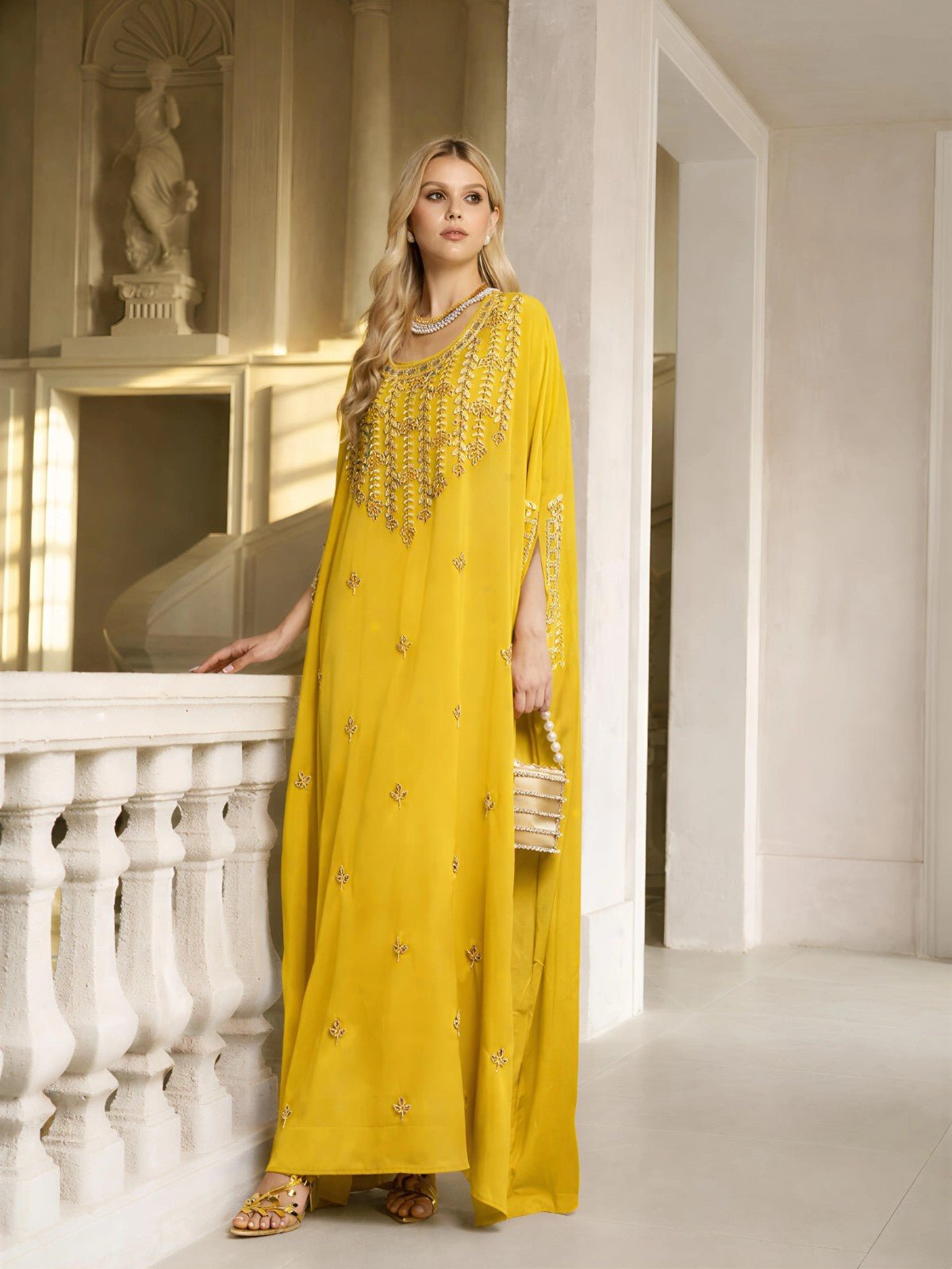 A woman stands indoors by a white railing in the Gilded Leaf Embellished Cape Dress by Social Queen, featuring yellow fabric, cape-style slit sleeves, and delicate embellishments, holding a small handbag as light streams in.