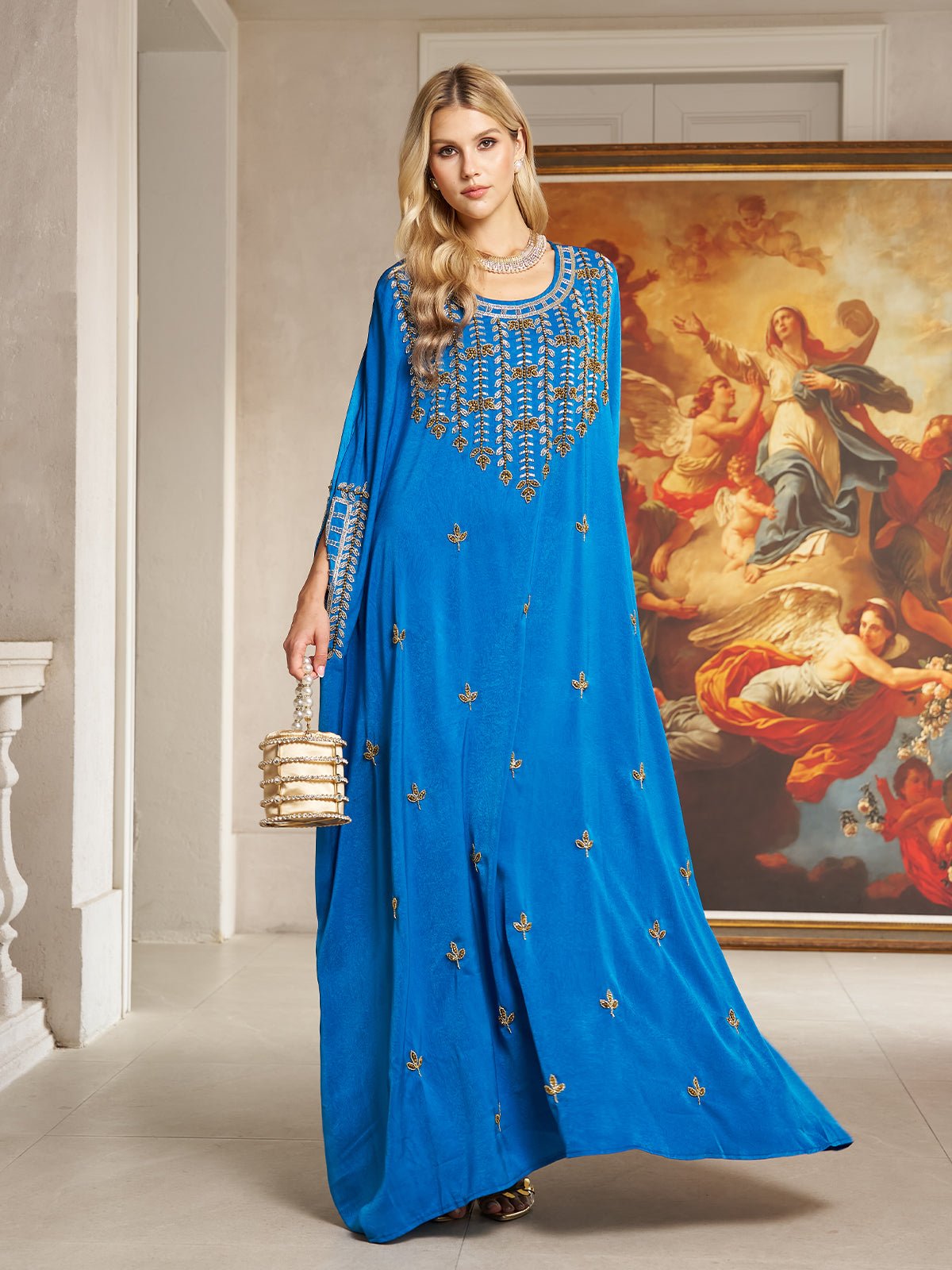 A woman wears the Gilded Leaf Embellished Cape Dress by Social Queen, a royal blue chiffon satin gown with cape-style slit sleeves, indoors with a gold handbag and a classical painting in the background.