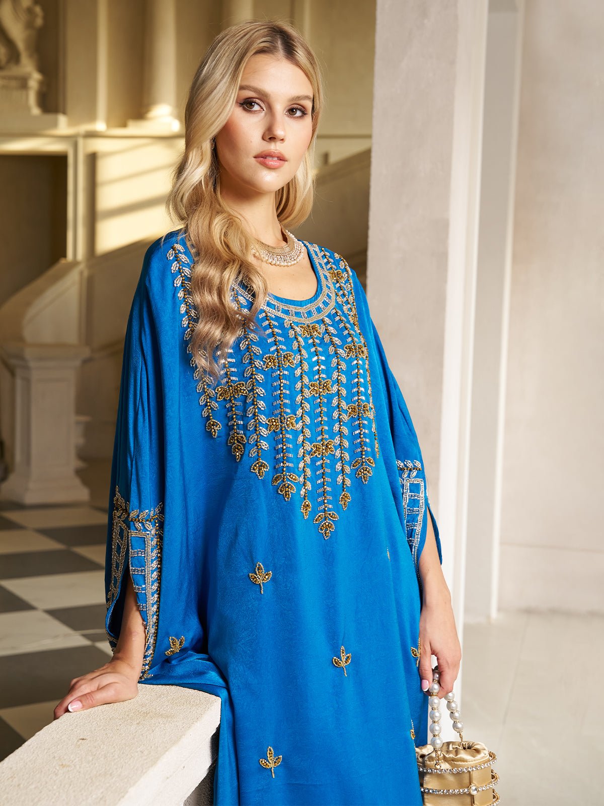 A woman stands indoors holding a white handbag, wearing the Social Queen Gilded Leaf Embellished Cape Dress—royal blue chiffon satin with gold leaf embroidery and cape-style slit sleeves. She has long blonde hair and a silver necklace.