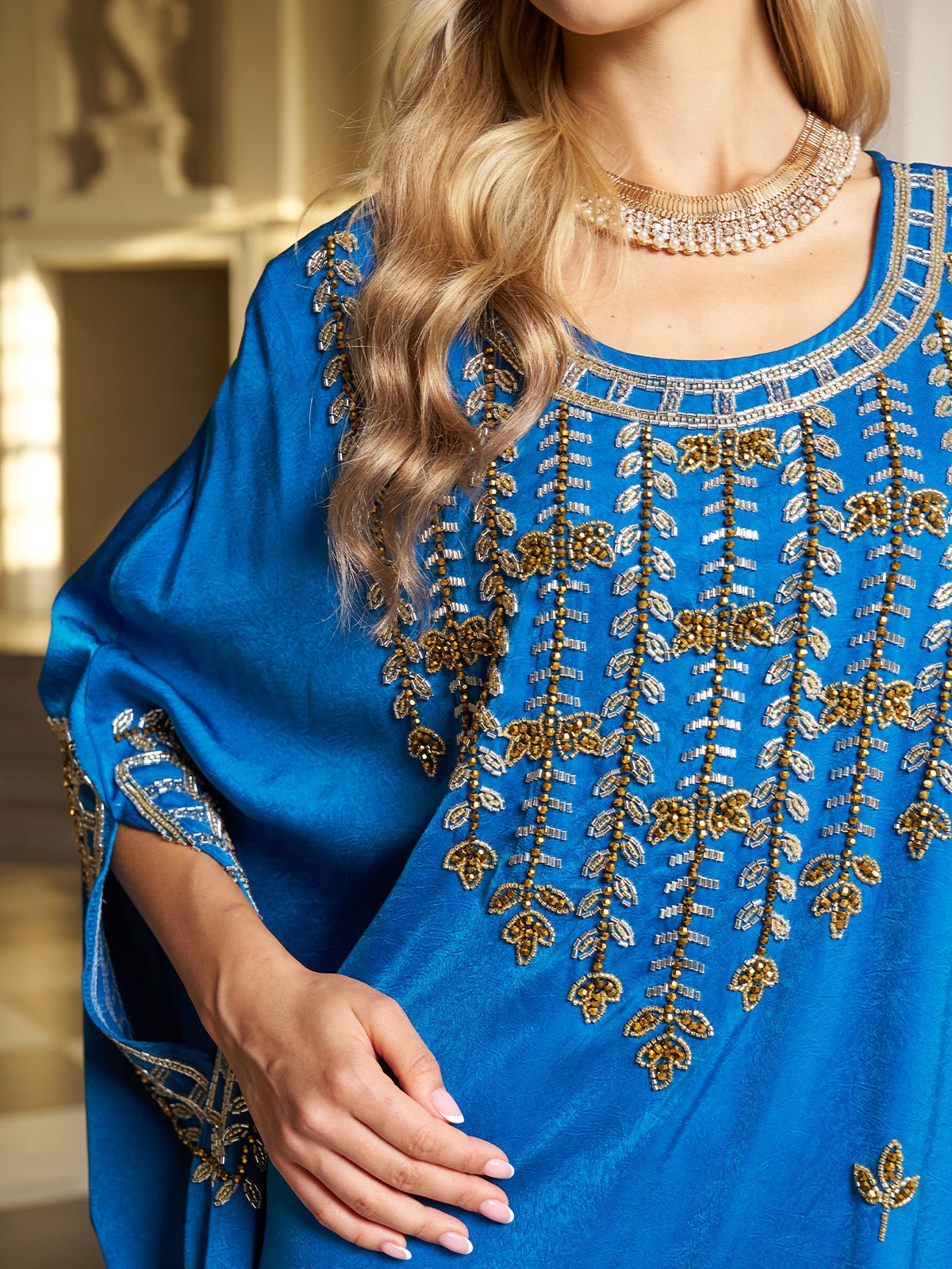 A woman wears the Social Queen Gilded Leaf Embellished Cape Dress in royal blue chiffon satin with gold and silver details, cape-style slit sleeves, and a layered necklace. She stands indoors with her face partially out of frame.