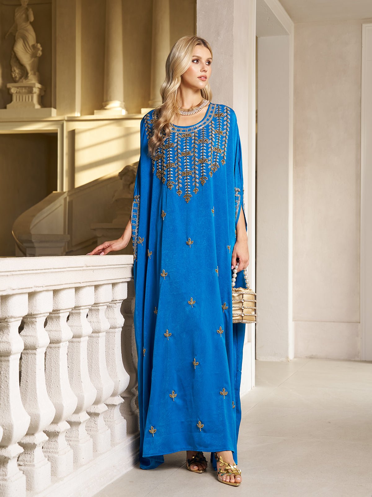 A woman wears the Social Queen Gilded Leaf Embellished Cape Dress in royal blue chiffon satin with gold leaf embroidery, cape-style slit sleeves, and pairs it with gold sandals and a small gold handbag by a white railing indoors.