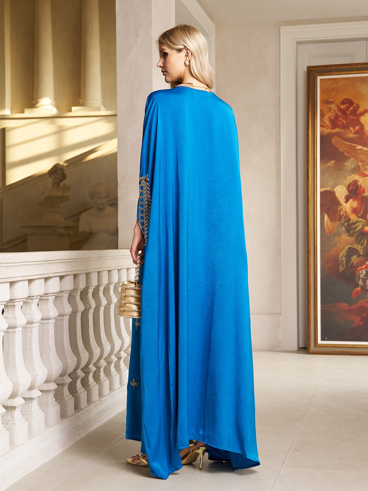 A woman stands on an indoor balcony beside a large framed painting, wearing the Social Queen Gilded Leaf Embellished Cape Dress in royal blue chiffon satin and holding a gold purse.
