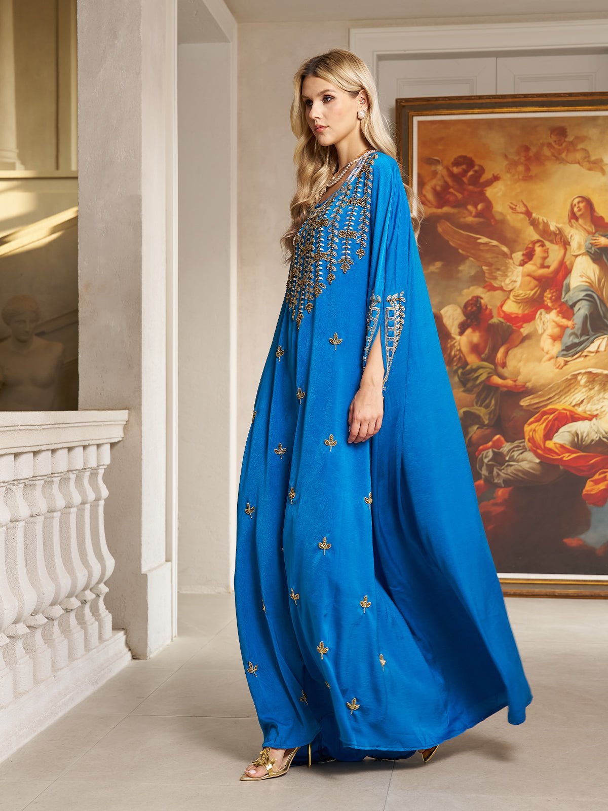 A woman in the Social Queen Gilded Leaf Embellished Cape Dress poses indoors near a painting, with light walls and ornate railings providing an elegant backdrop.