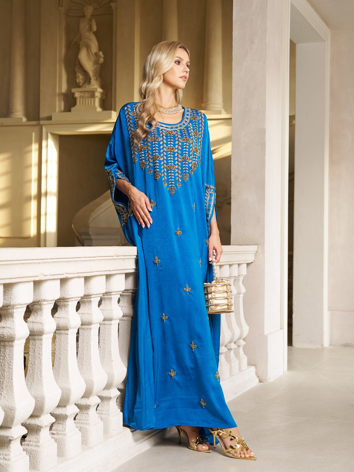 A woman stands indoors by a white balustrade, wearing the Social Queen Gilded Leaf Embellished Cape Dress in royal blue chiffon satin, with slit cape sleeves, holding a small handbag and looking to the side.