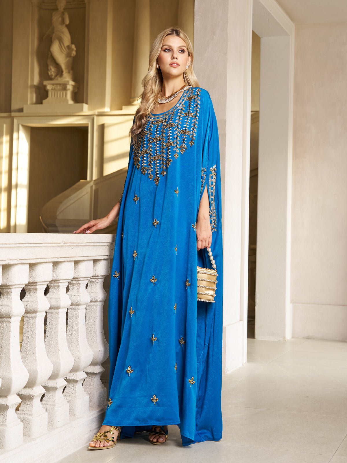 A woman wearing the Social Queen Gilded Leaf Embellished Cape Dress stands indoors by a white balustrade, holding a small handbag and pairing her look with gold sandals.