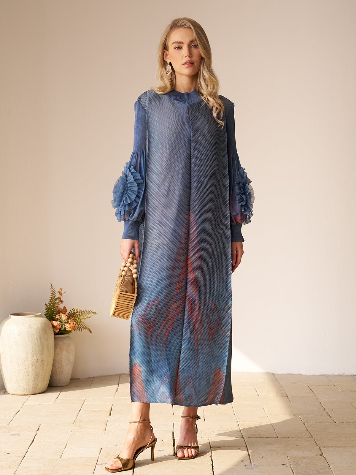 Indoors, a woman wears the Social Queen Gradient Pleated Dress with 3D Floral Sleeves—ideal for a wedding guest. She accessorizes with a wicker handbag and heeled sandals.
