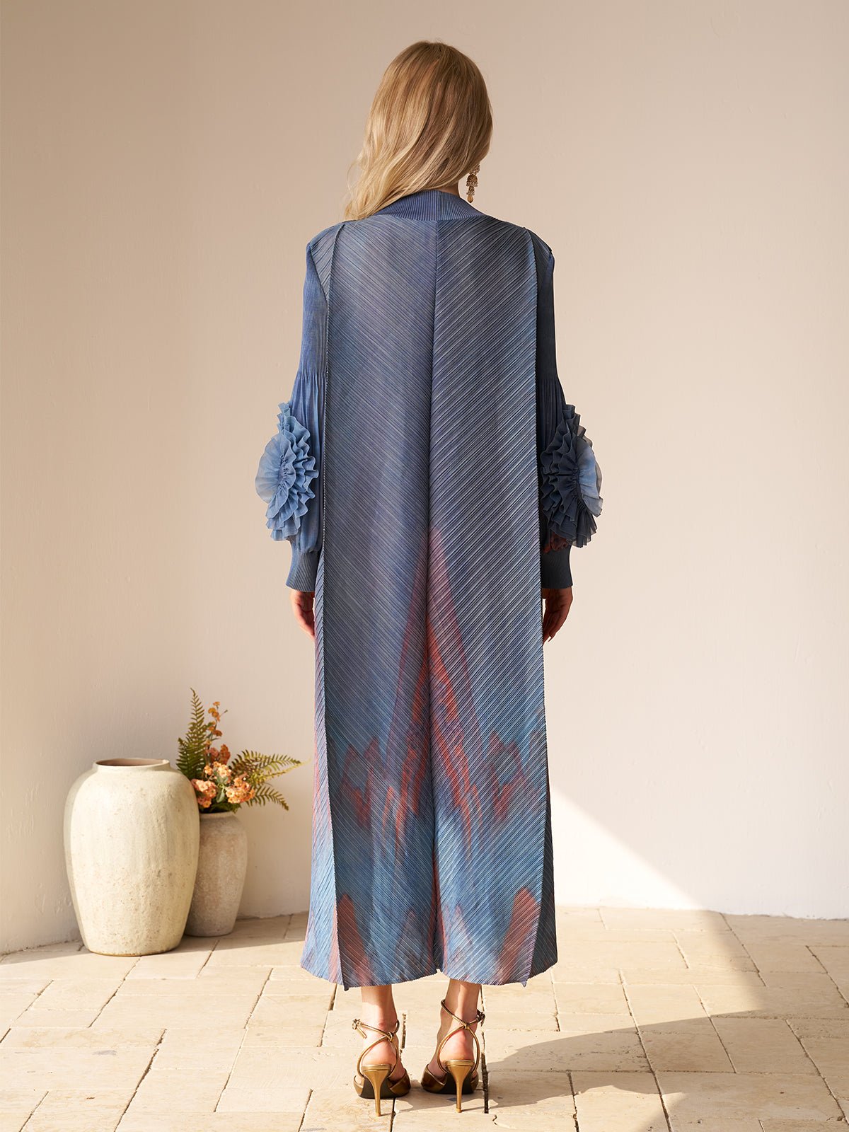 A woman stands indoors facing away, wearing Social Queen's Gradient Pleated Dress with 3D Floral Sleeves and heels—ideal for a wedding guest. Two ceramic vases with flowers sit to her left on the floor.