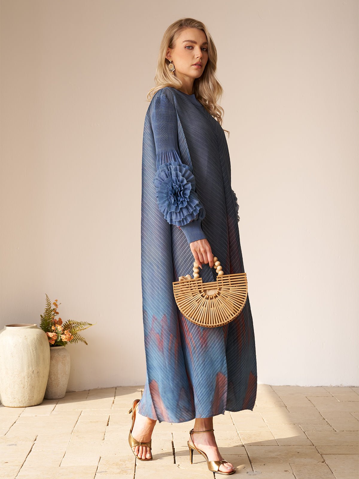 A woman stands indoors by decorative vases, wearing the Social Queen Gradient Pleated Dress with 3D Floral Sleeves, accessorized with a bamboo handbag and gold heels—an ideal choice for a wedding guest.