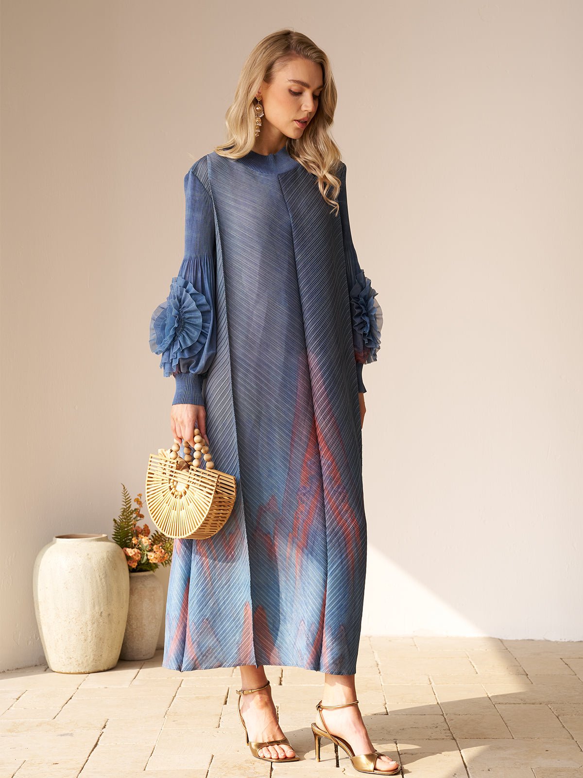 A woman indoors holds a wooden handbag and wears gold heels with the Social Queen Gradient Pleated Dress with 3D Floral Sleeves. Two ceramic vases with plants are behind her—an ideal, stylish look for a wedding guest.