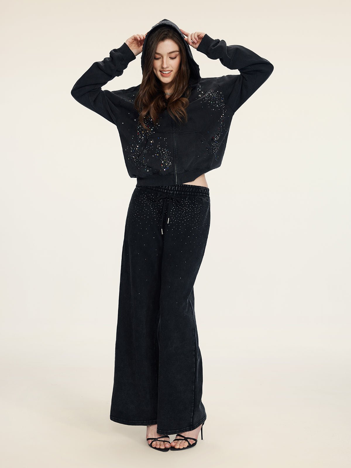 A woman faces forward in the Social Queen Hand-Applied Rhinestone Washed Hoodie with matching wide-leg pants, finished with black heeled sandals.