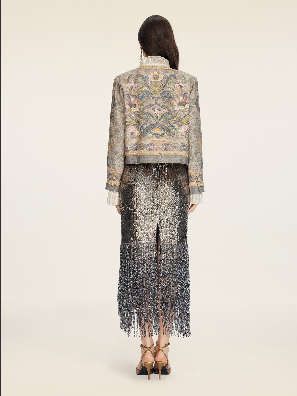 A person stands facing away on a plain light background, wearing the Hand-Beaded Printed Jacket by Social Queen paired with a metallic, fringed skirt and high heels.