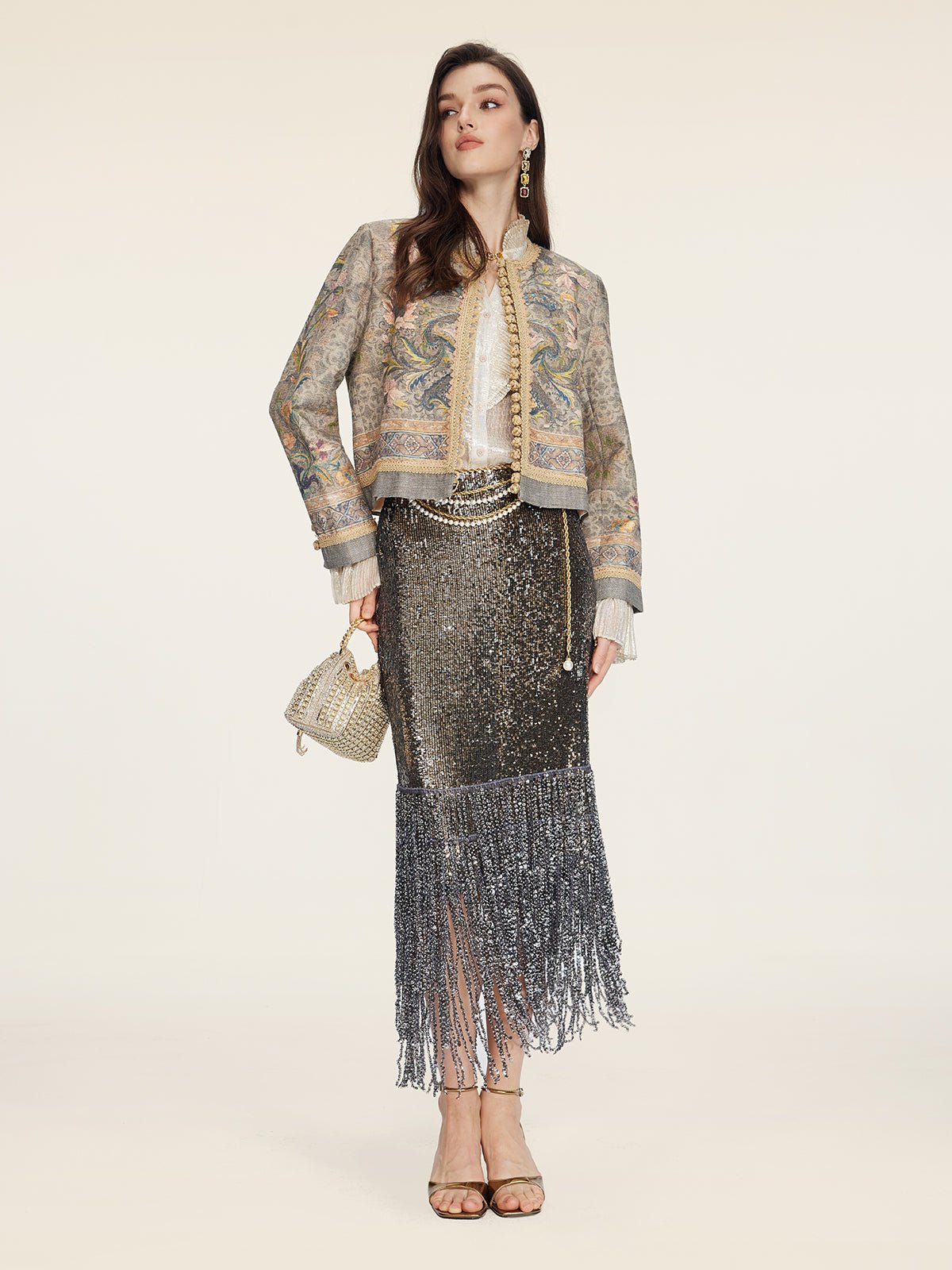A woman stands against a plain background wearing the Social Queen Hand-Beaded Printed Jacket, paired with a white blouse, metallic fringe skirt, gold heels, and a small gold handbag.