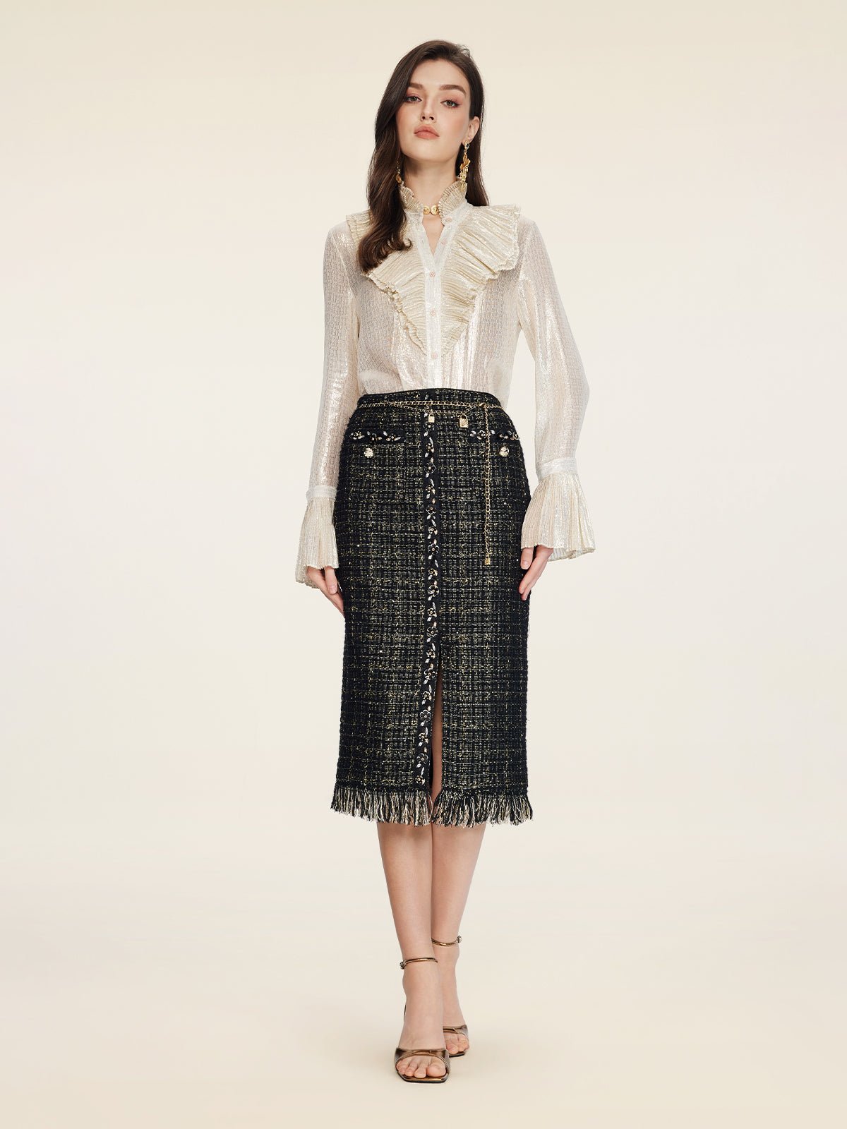 A woman faces forward wearing a sheer white ruffled blouse and the Social Queen Hand-Beaded Tweed Fringe Slit Skirt, featuring black-and-gold details, front buttons, a fringe hem, and paired with strappy heeled sandals.