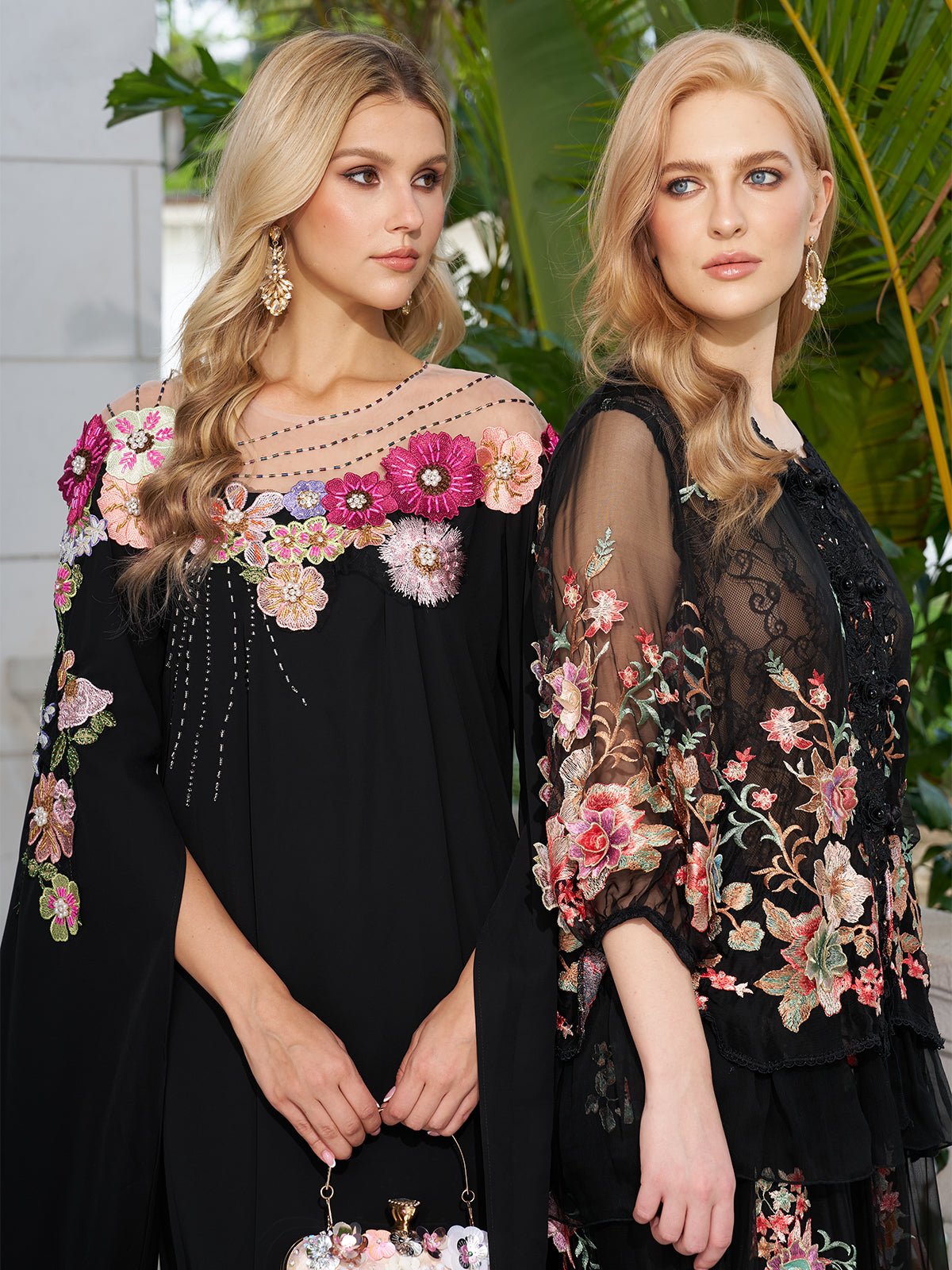 Two women in black Social Queen Hand-Embroidered Floral Cape Gowns stand outdoors amid lush greenery, their elegant gowns making a perfect choice for wedding guests.