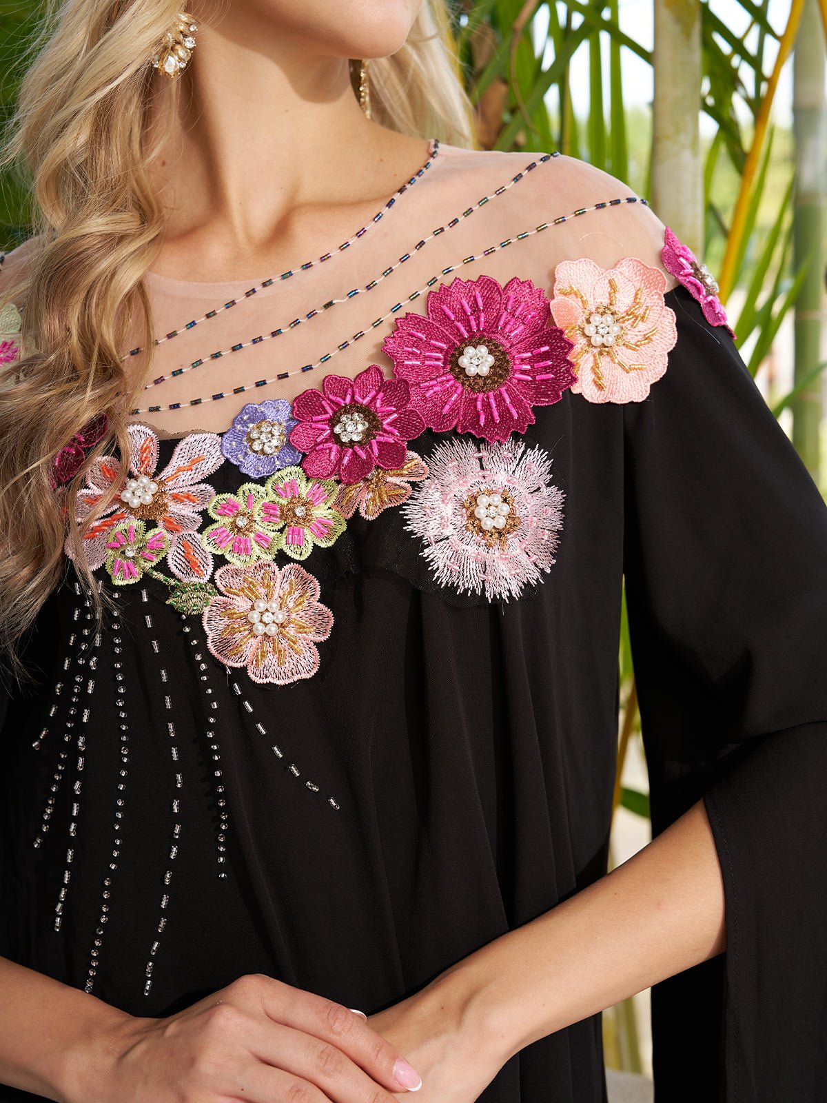 A woman wears the Social Queen Hand-Embroidered Floral Cape Gown—black with a sheer neckline and colorful floral appliqués—making it an elegant wedding guest dress.