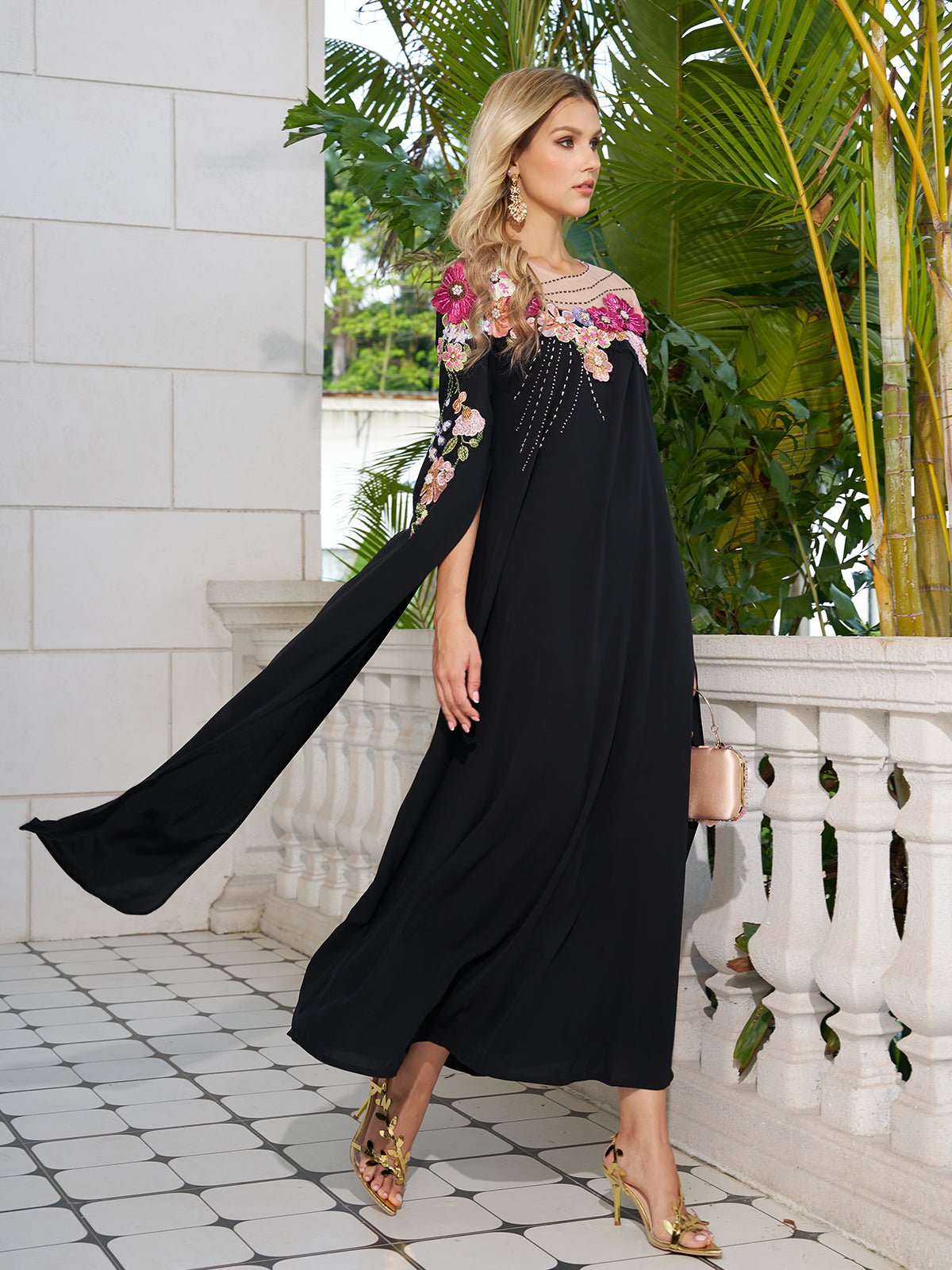 A woman in the Social Queen Hand-Embroidered Floral Cape Gown walks on a tiled balcony, clutch in hand, surrounded by lush green plants and a white stone railing.